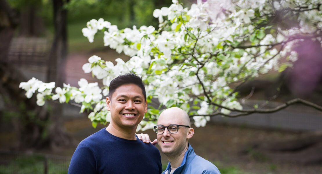 Two men standing in front of tree with white flowers from a Central Park engagement photo shoot