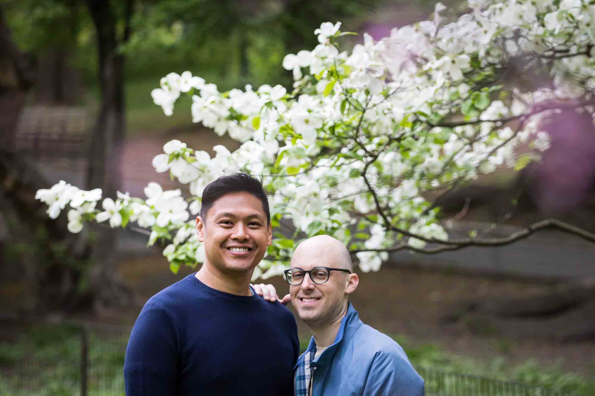 Two men standing in front of tree with white flowers from a Central Park engagement photo shoot