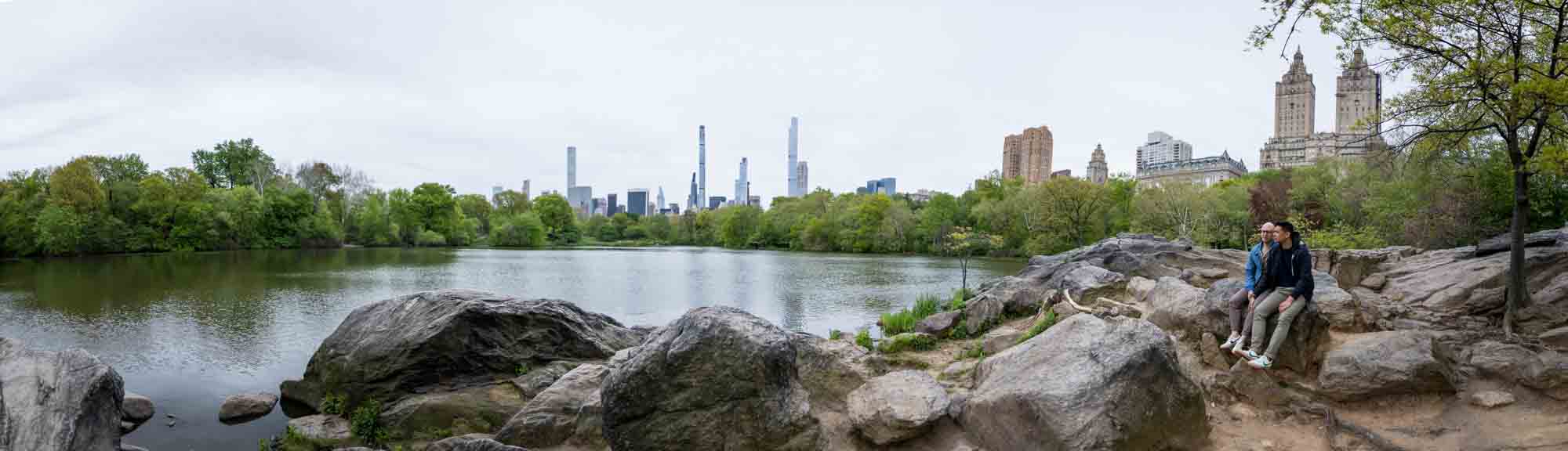 Panoramic view of two men sitting on rock in front of Central Park Lake
