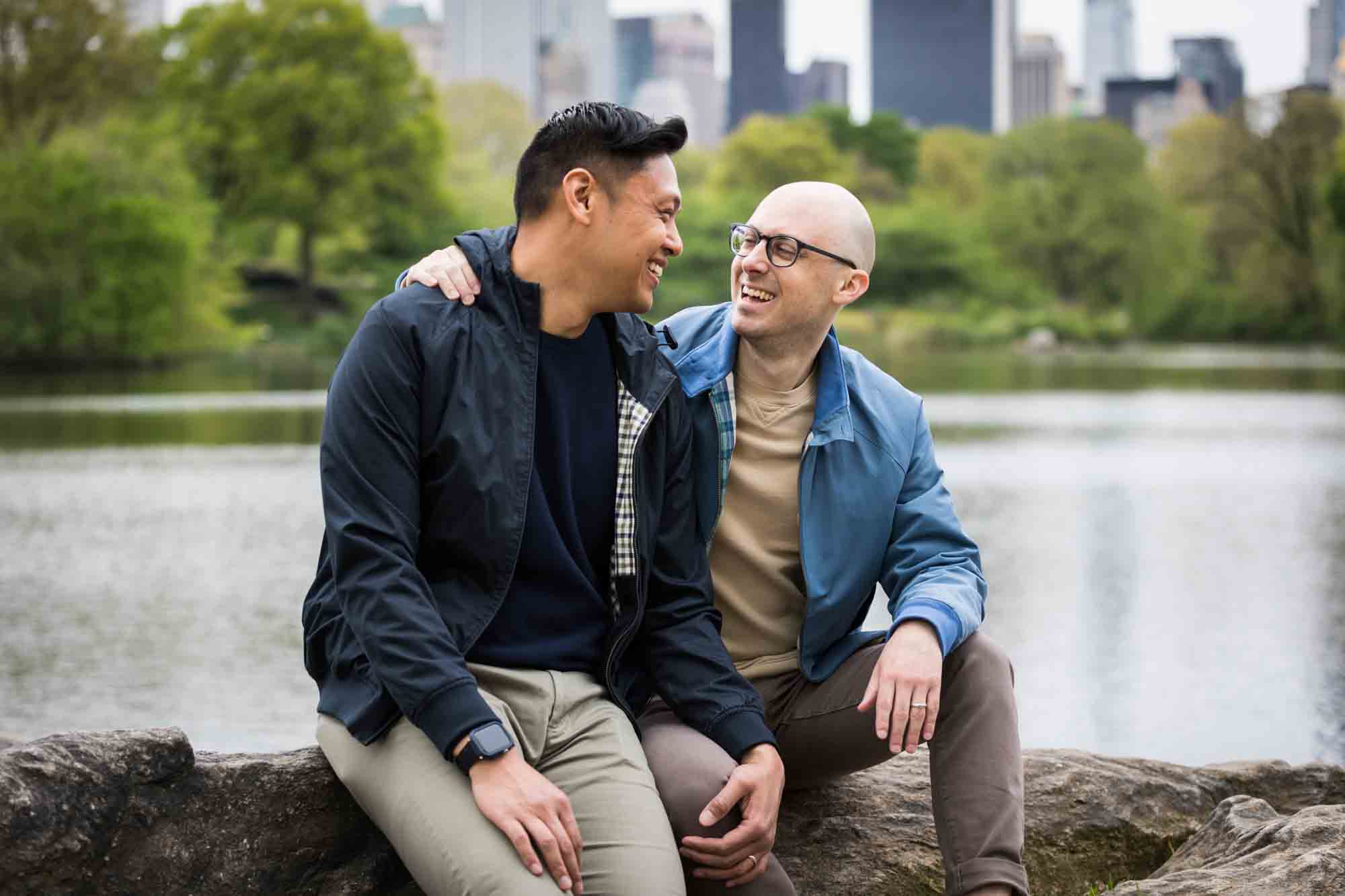 Two men sitting on a rock with NYC skyline in background from a Central Park engagement photo shoot