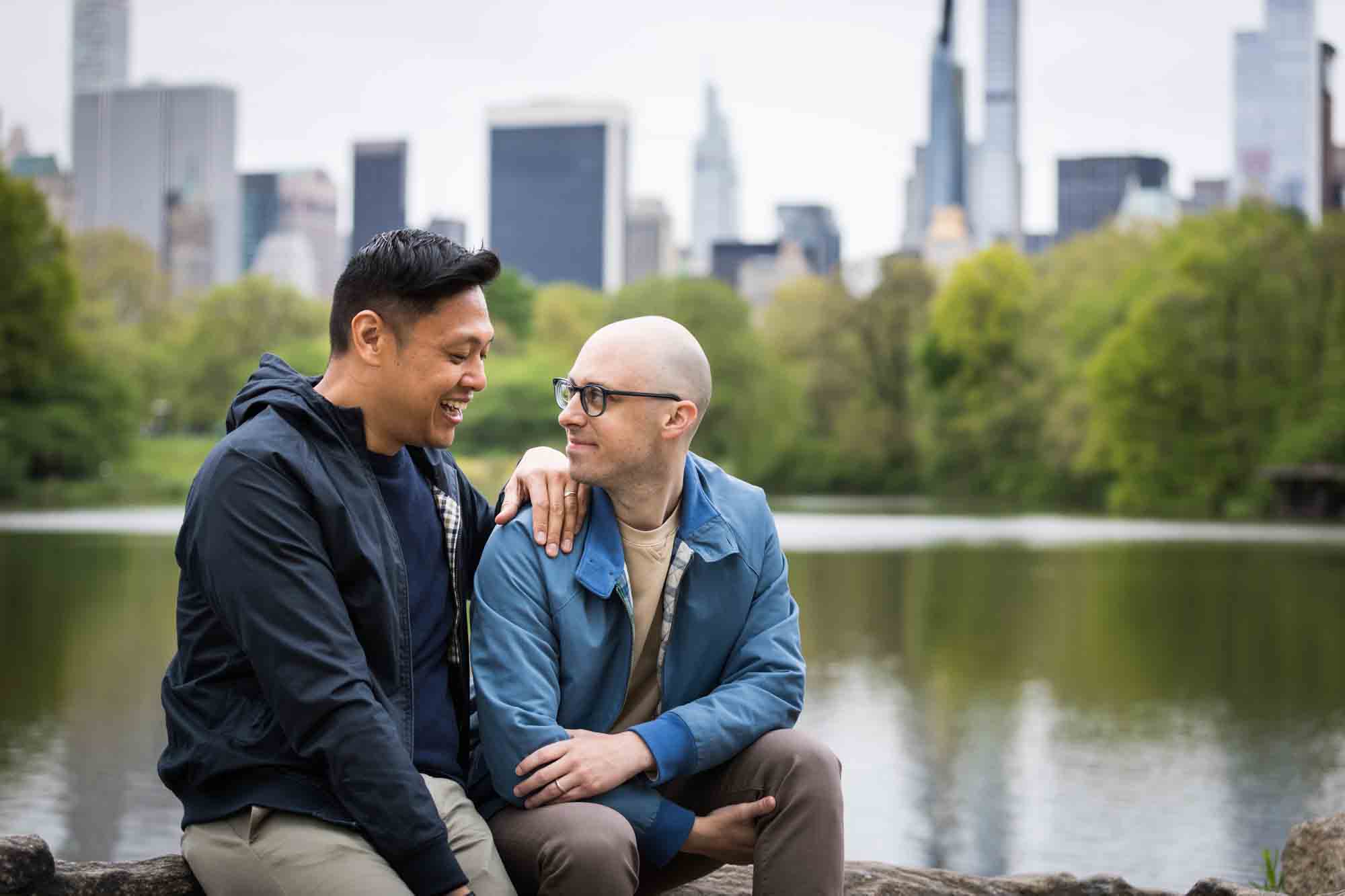 Two men sitting on rock in front of Central Park Lake with NYC skyline in background