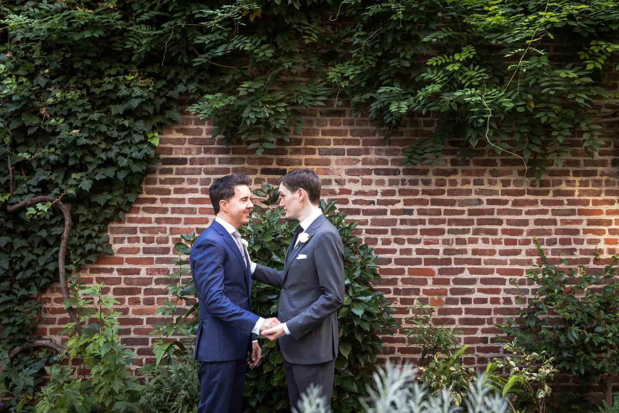 Two grooms holding hands in front of a brick wall for article on wedding bouquet photo tips
