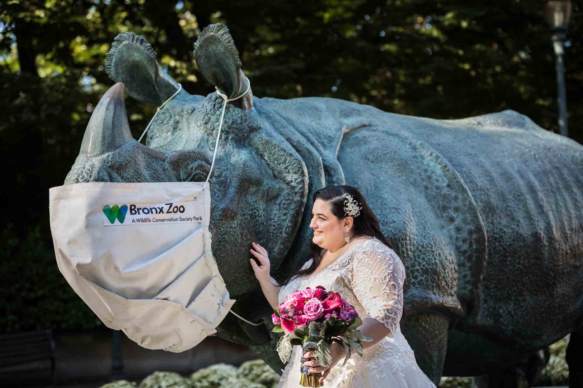 Bride holding pink bouquet and touching rhino statue wearing large cloth mask