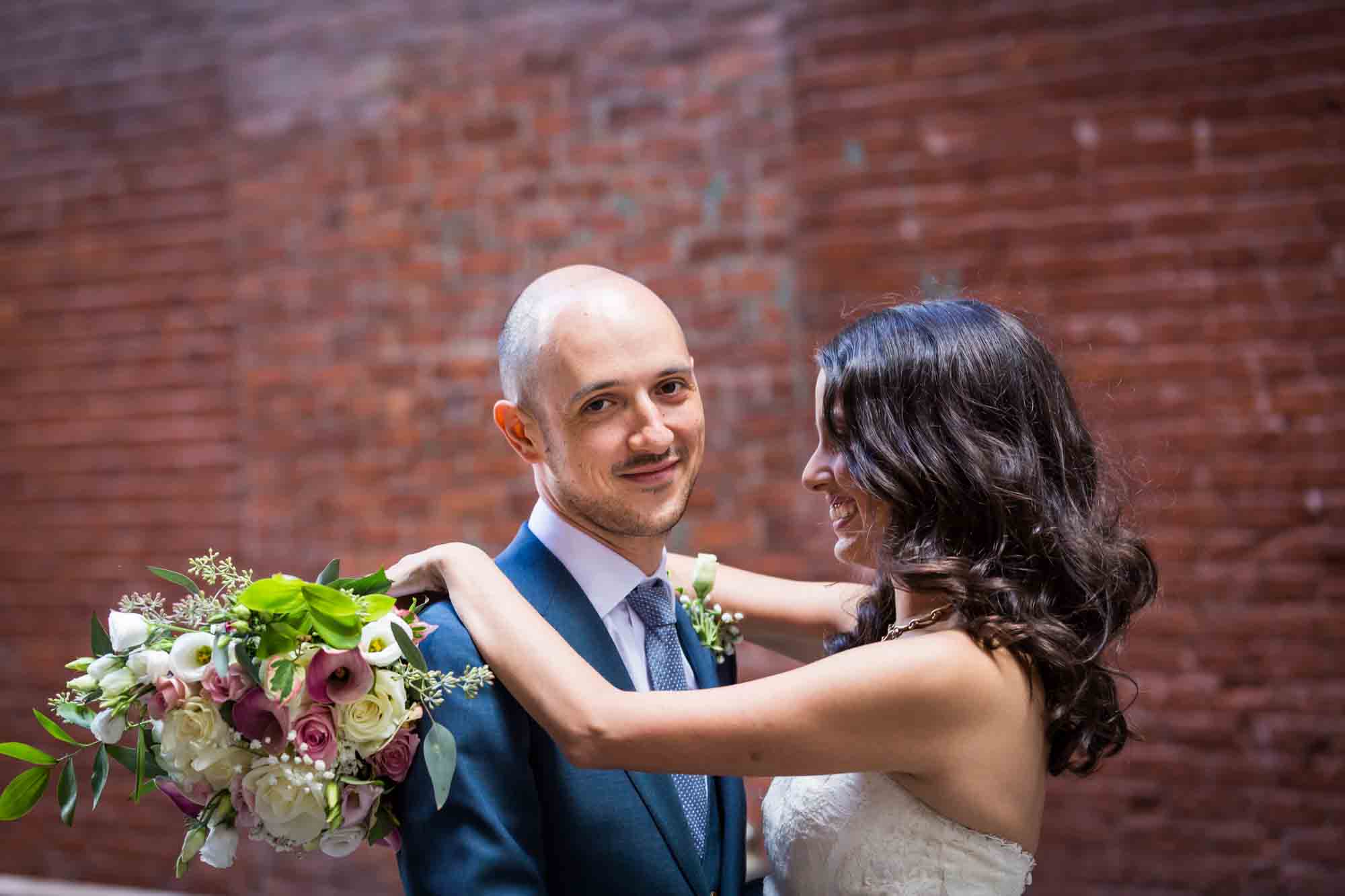 Bride holding bouquet and hugging groom against brick wall