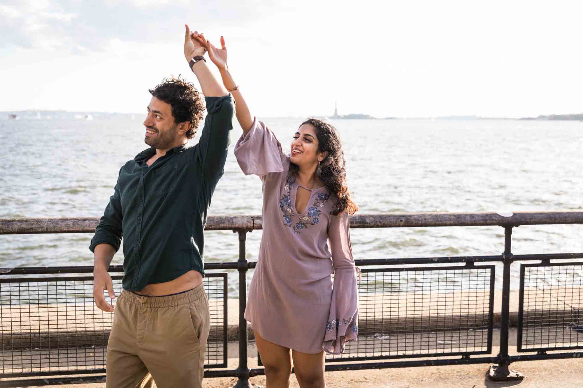 Battery Park engagement photos of couple dancing in front of NYC waterfront with hands raised