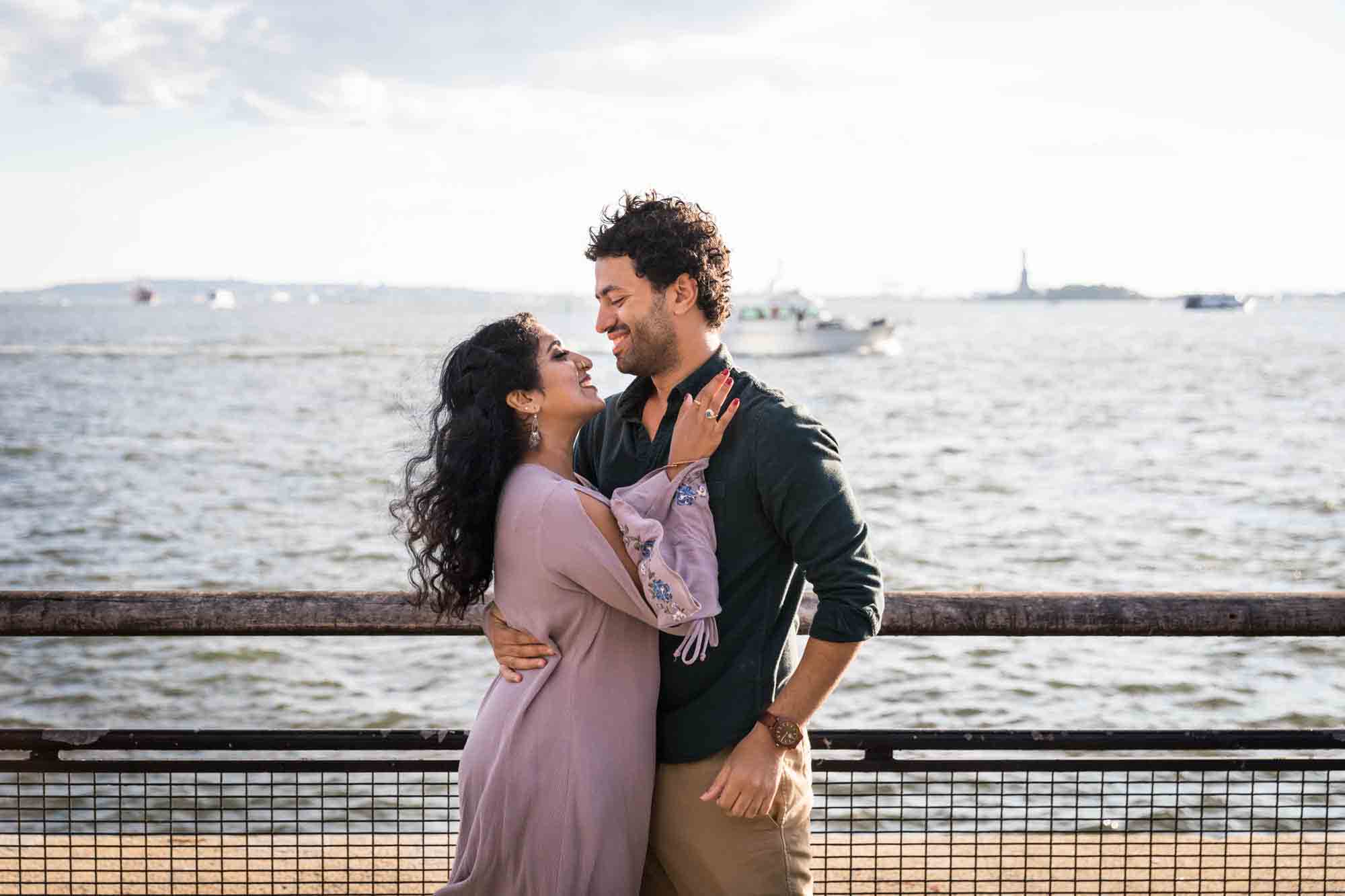 Battery Park engagement photos of couple hugging in front of railing and NYC waterfront