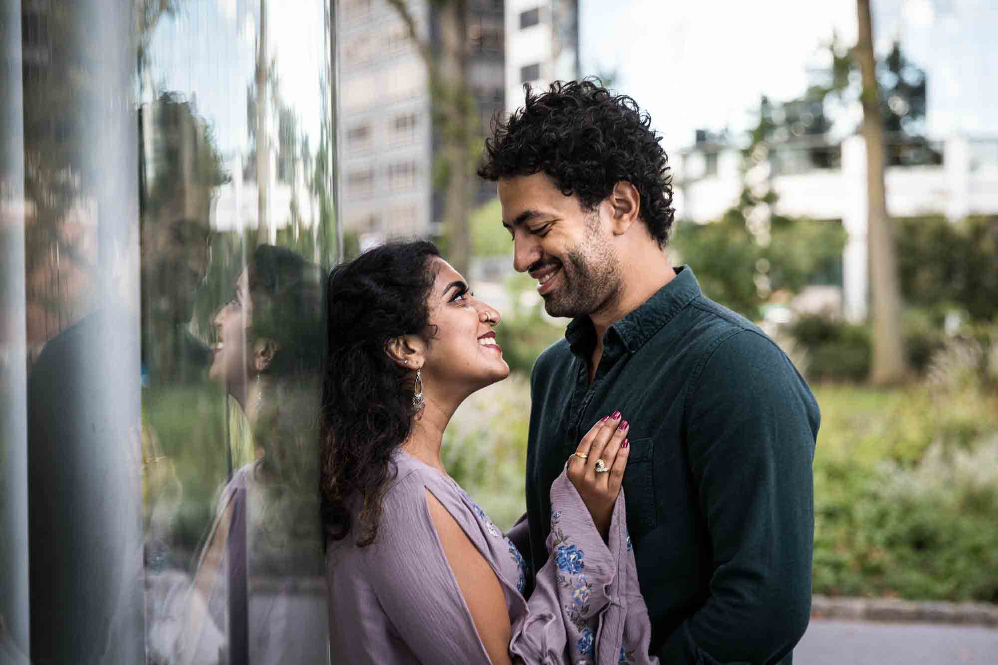 Battery Park engagement photos of couple leaning against SeaGlass Carousel with reflection