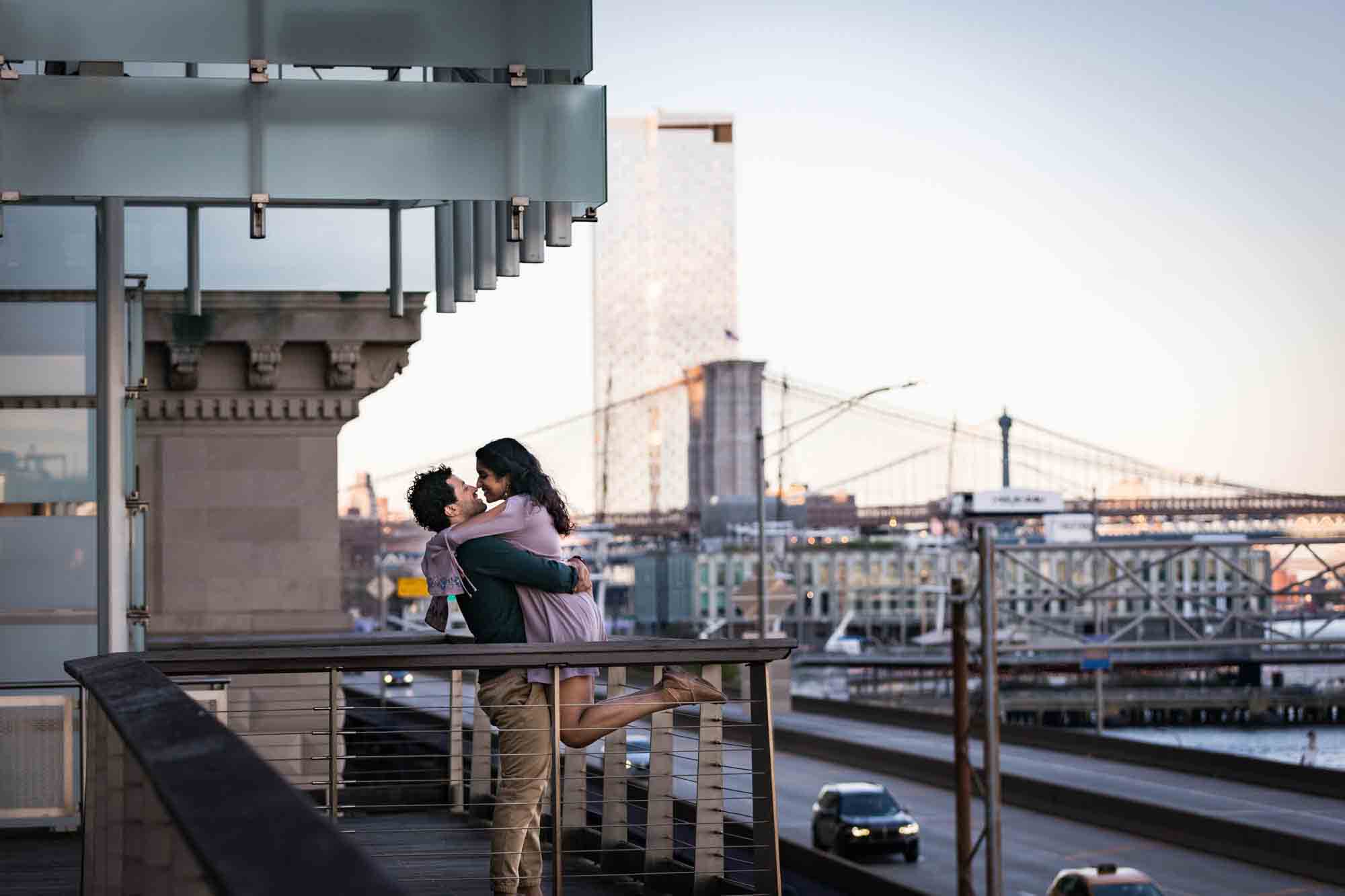 Man lifting up woman on pathway at Elevated Acre with Brooklyn Bridge in background