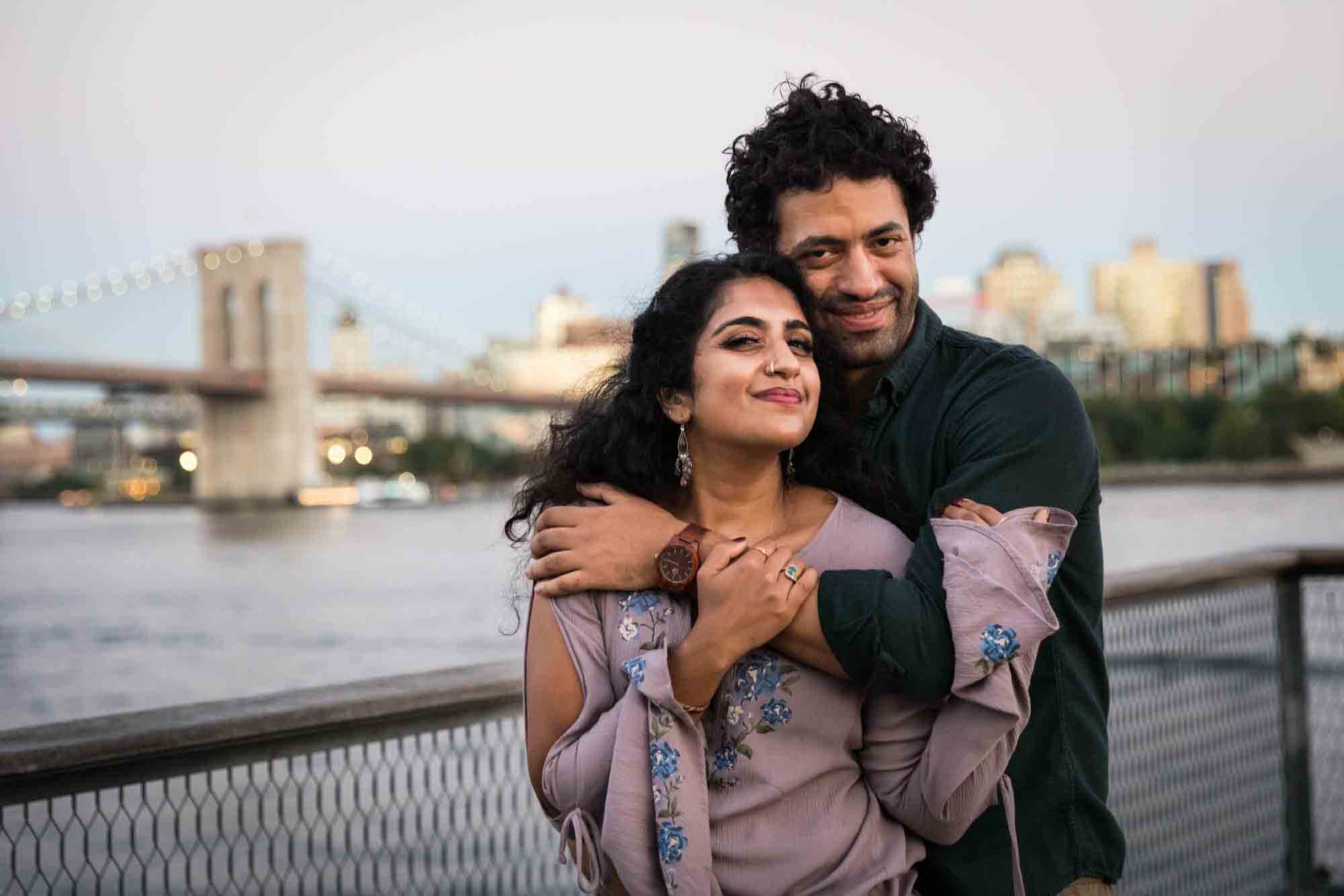 Couple hugging on Pier 17 with Brooklyn Bridge at sunset in background