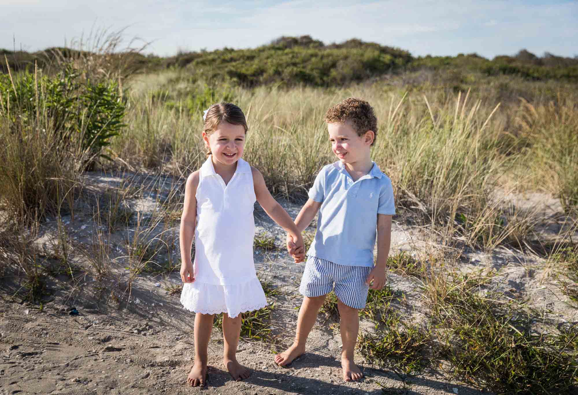 Two children holding hands on a beach in front of dunes for an article on beach family portrait tips