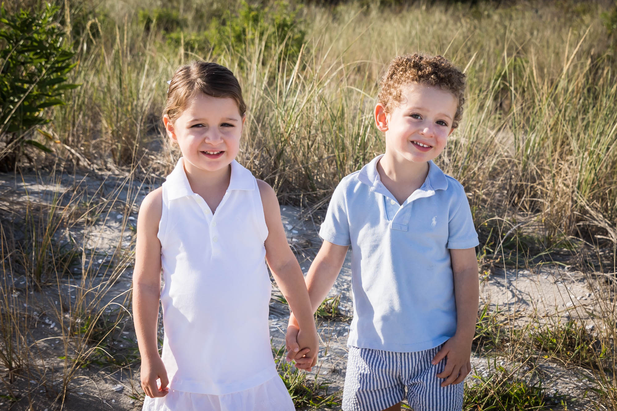 Two children holding hands on a beach in front of dunes for an article on beach family portrait tips