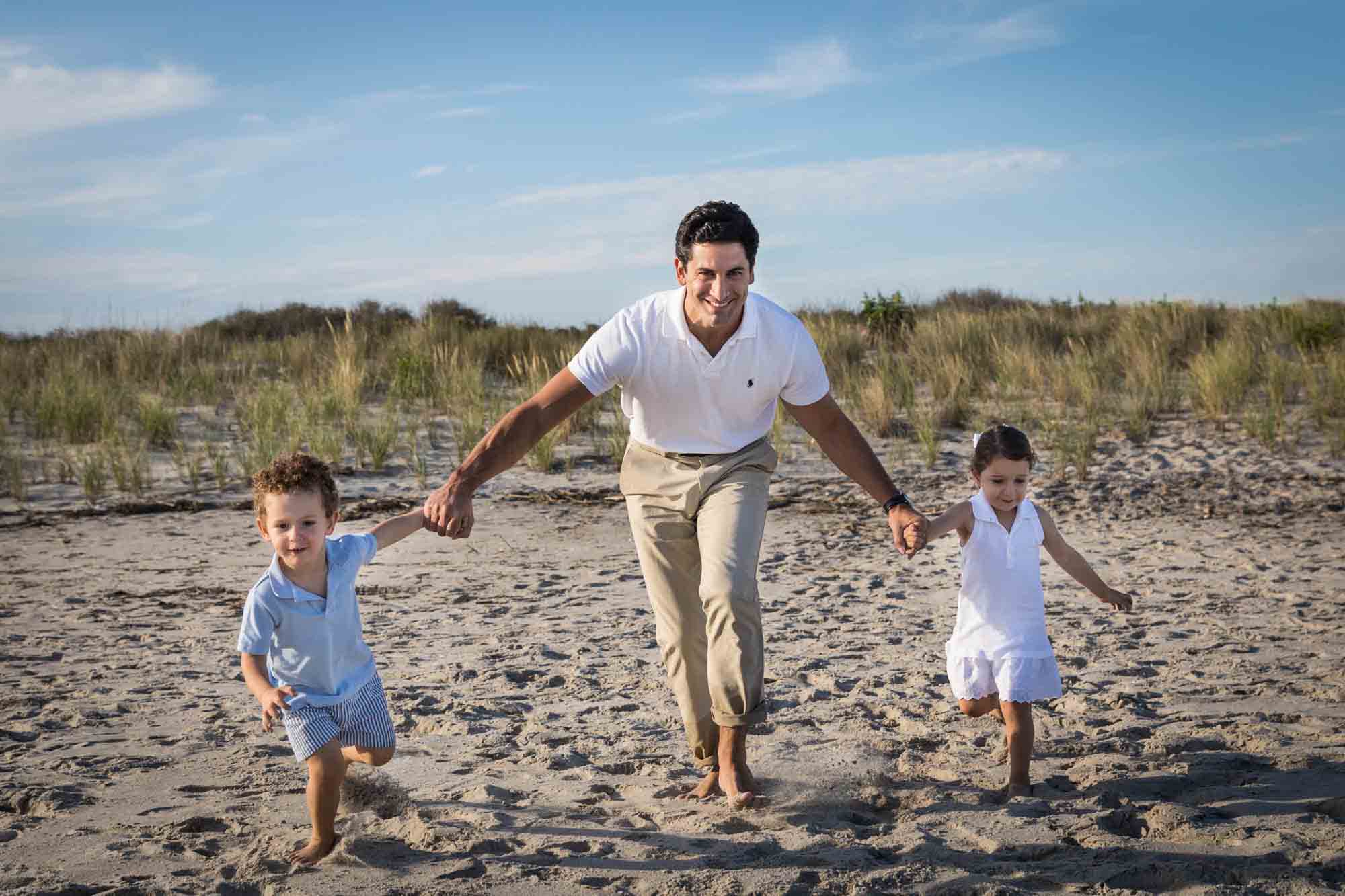 Father holding hands of two small children while running on beach for an article on beach family portrait tips