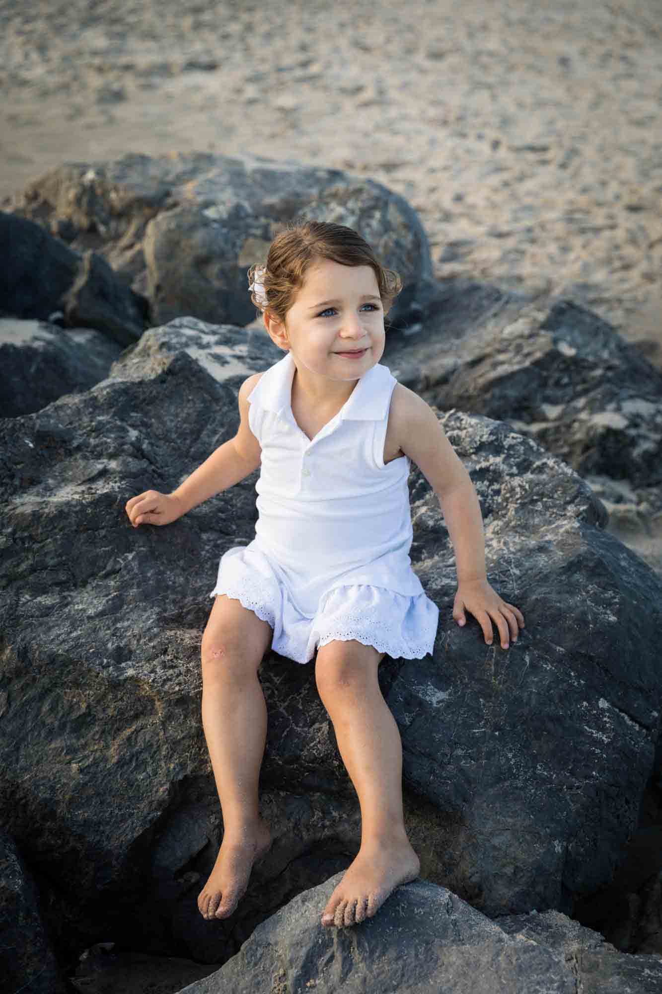 Beach family portrait of little girl in white dress sitting on rocks