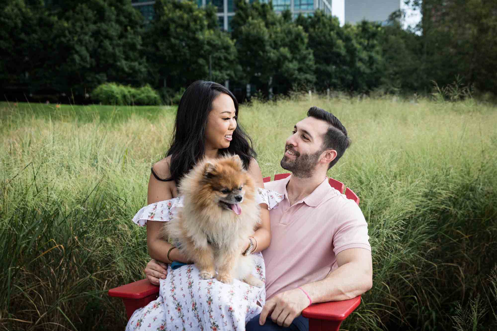 Couple sitting on red Adirondack chair with Pomeranian during a Gantry Plaza State Park engagement photo shoot