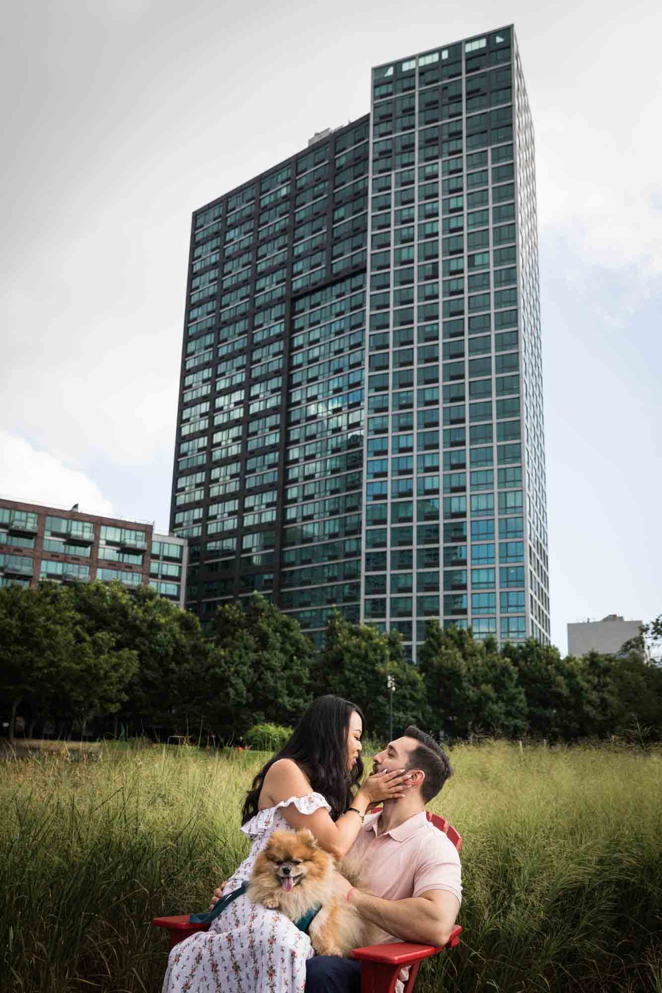 Couple sitting on red Adirondack chair kissing during a Gantry Plaza State Park engagement photo shoot