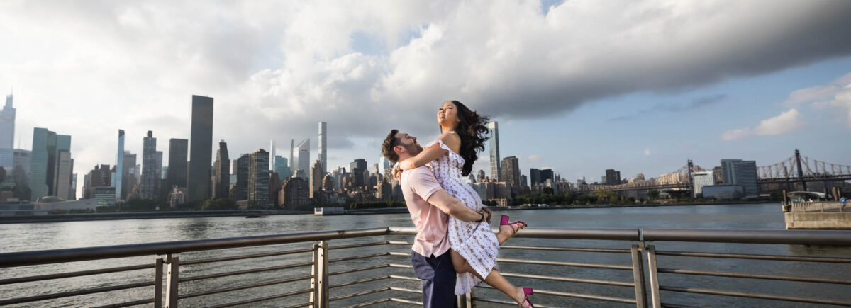 Man lifting up woman on boardwalk during Gantry Plaza State Park engagement session for an article on engagement portrait clothing tips