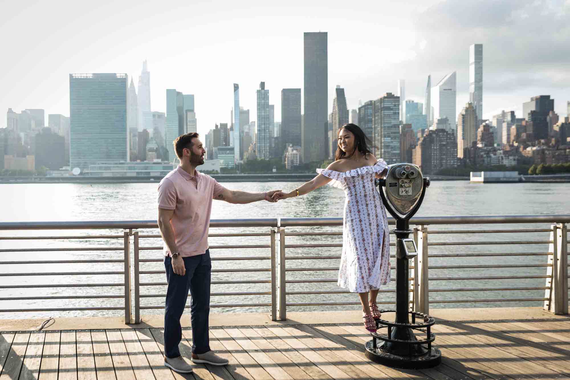 Couple holding hands and woman standing on viewfinder during a Gantry Plaza State Park engagement photo shoot