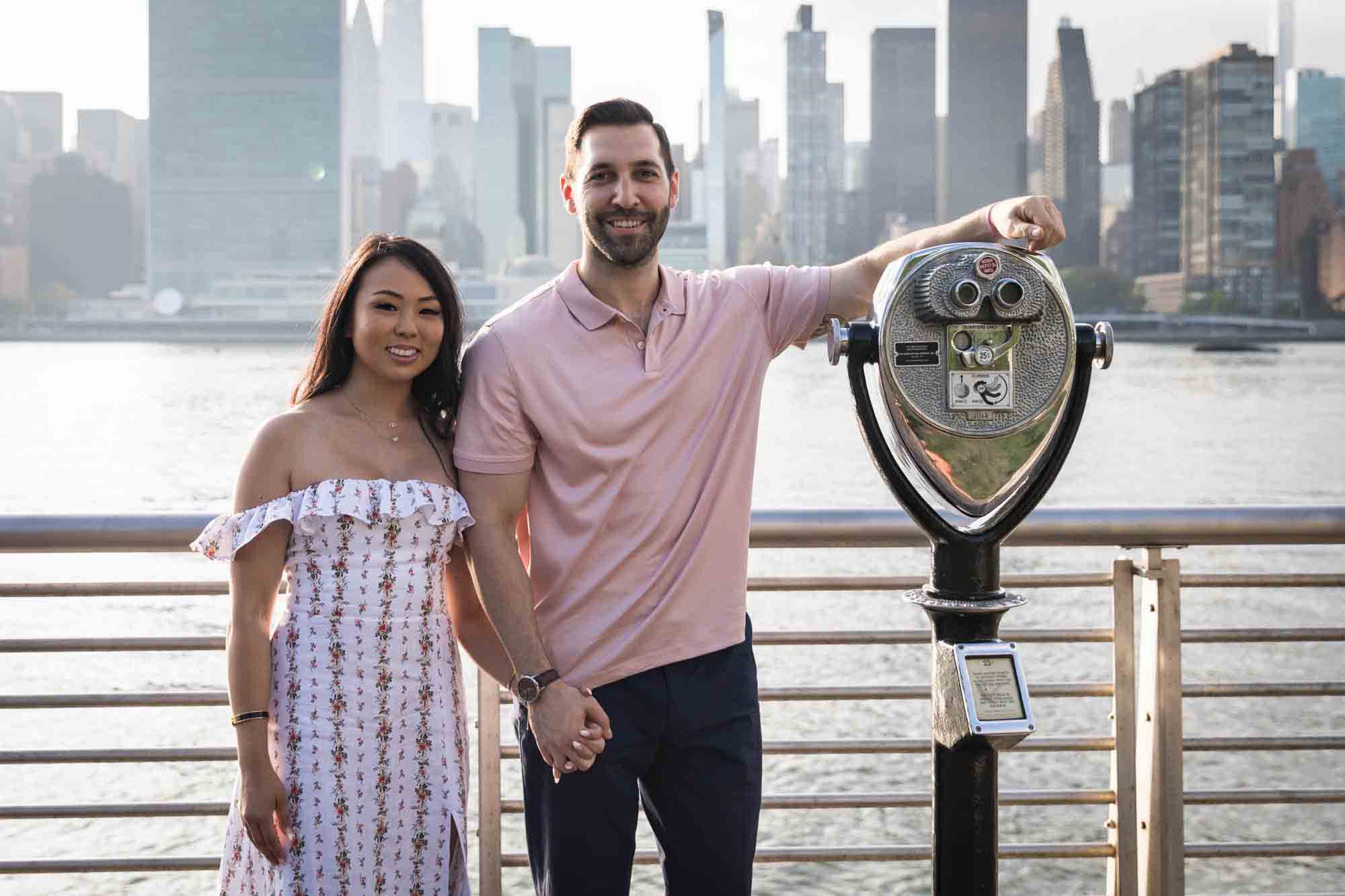 Couple on boardwalk with man leaning on viewfinder during Gantry Plaza State Park engagement session for an article on engagement portrait clothing tips