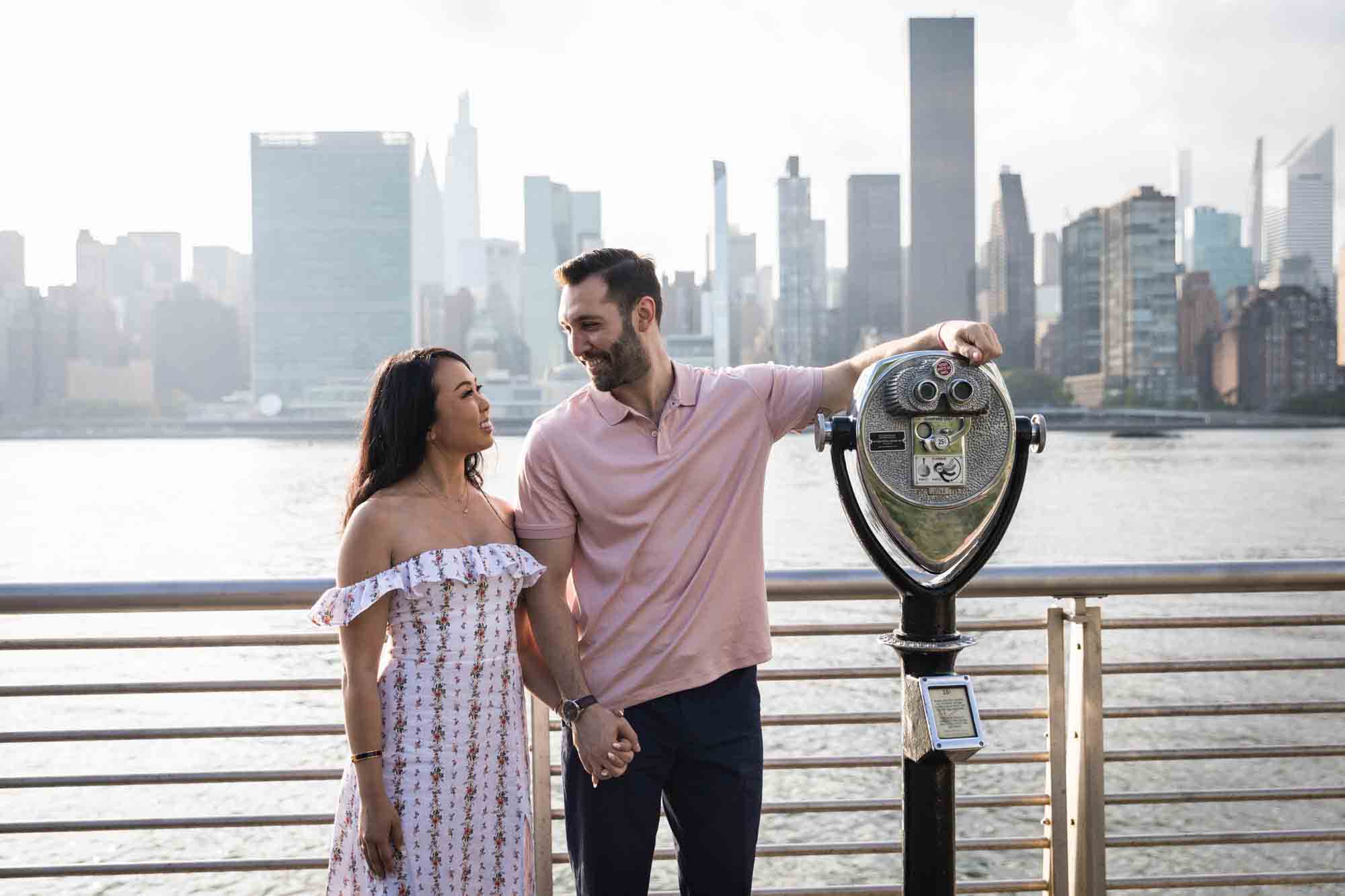 Couple standing on boardwalk and man leaning on viewfinder during a Gantry Plaza State Park engagement photo shoot