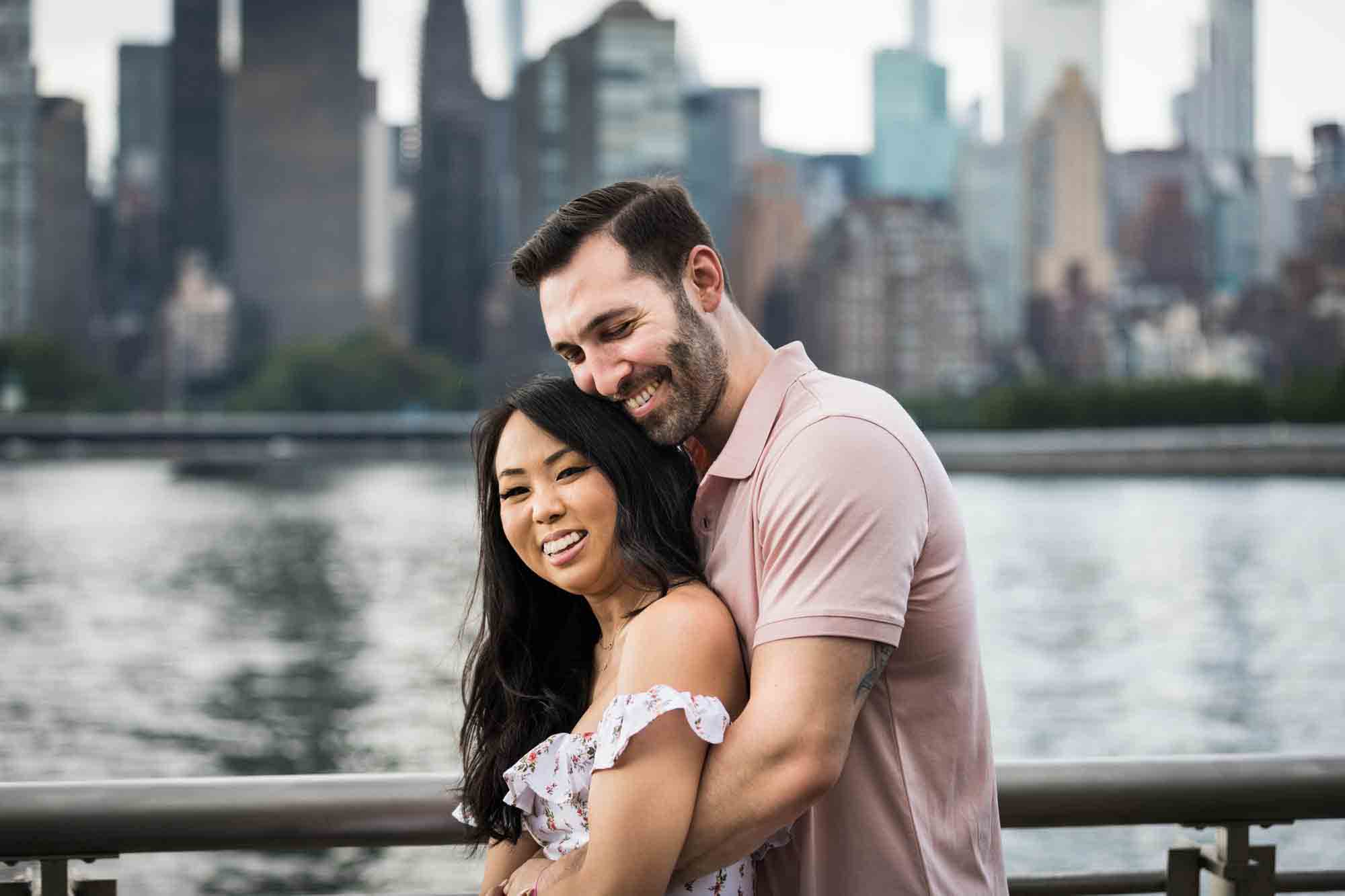 Couple hugging in front of NYC waterfront at Gantry Plaza State Park engagement session for an article on engagement portrait clothing tips