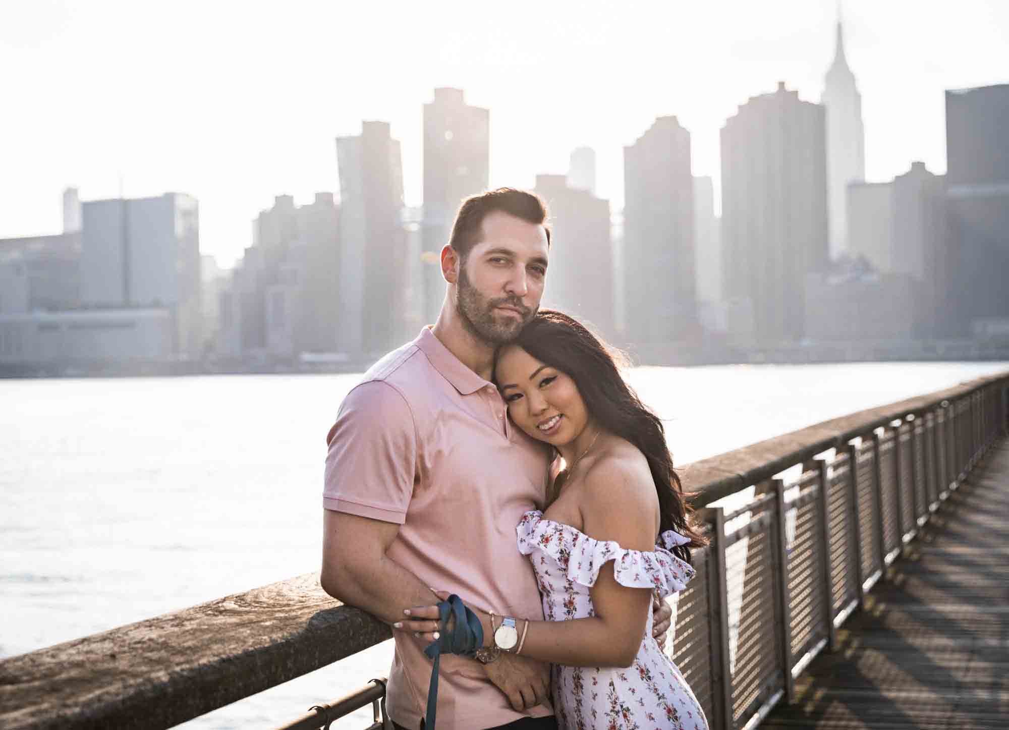 Couple leaning against railing with NYC skyline in background during a Gantry Plaza State Park engagement photo shoot