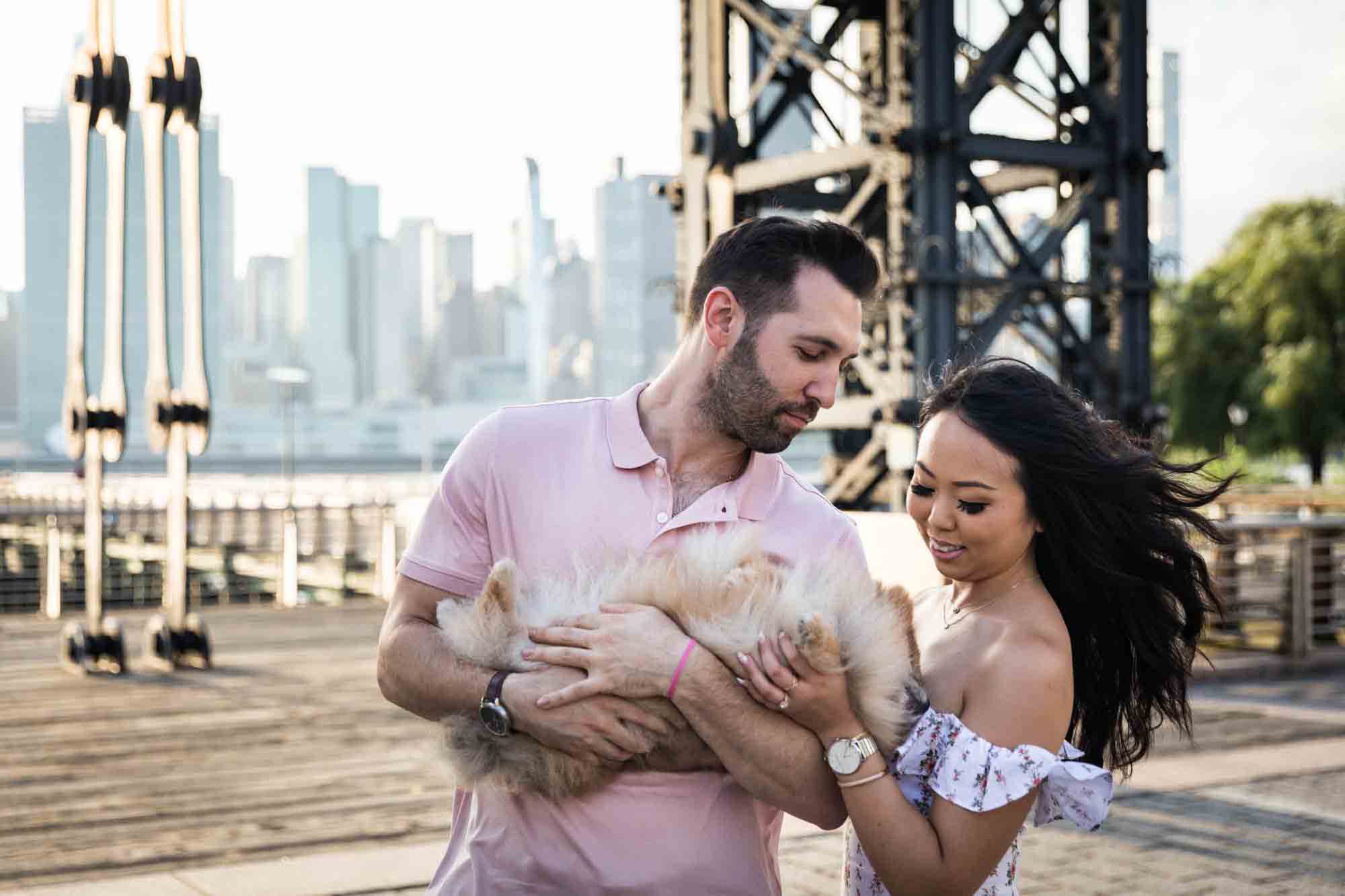 Couple petting Pomeranian in man's arms during Gantry Plaza State Park engagement session for an article on engagement portrait clothing tips