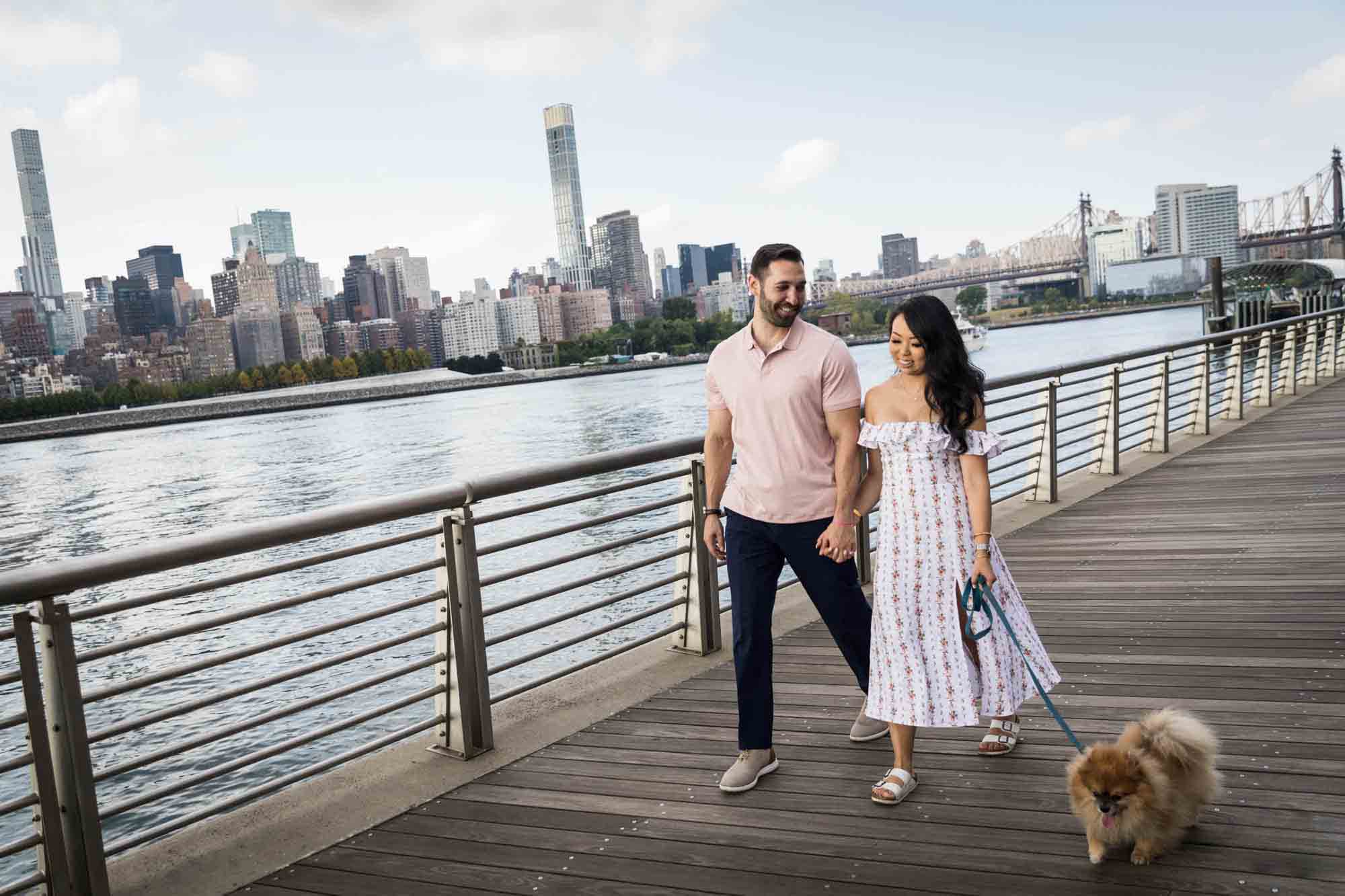 Couple walking on boardwalk with Pomeranian on leash during a Gantry Plaza State Park engagement photo shoot