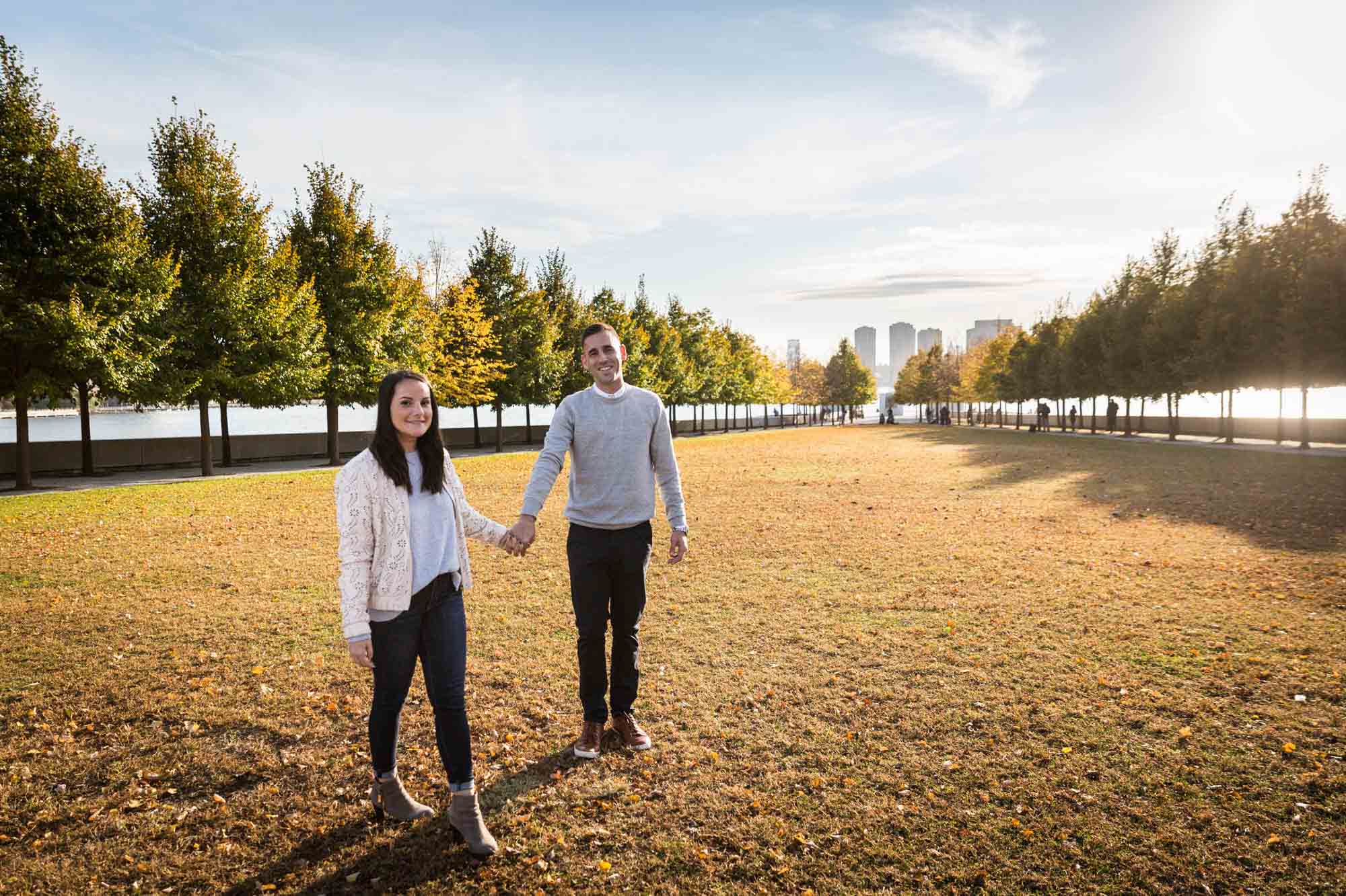 Couple holding hands and walking across grass during a Roosevelt Island engagement photo shoot