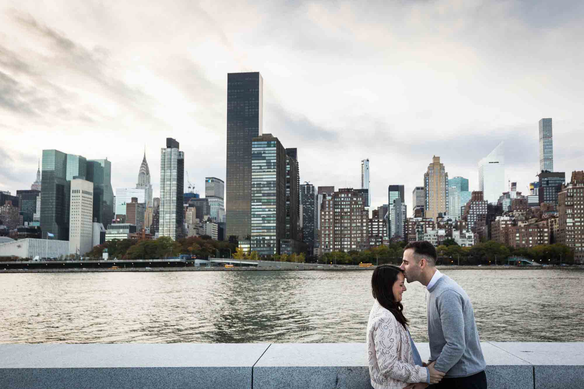 Man kissing woman on forehead in front of NYC skyline during a Roosevelt Island engagement photo shoot
