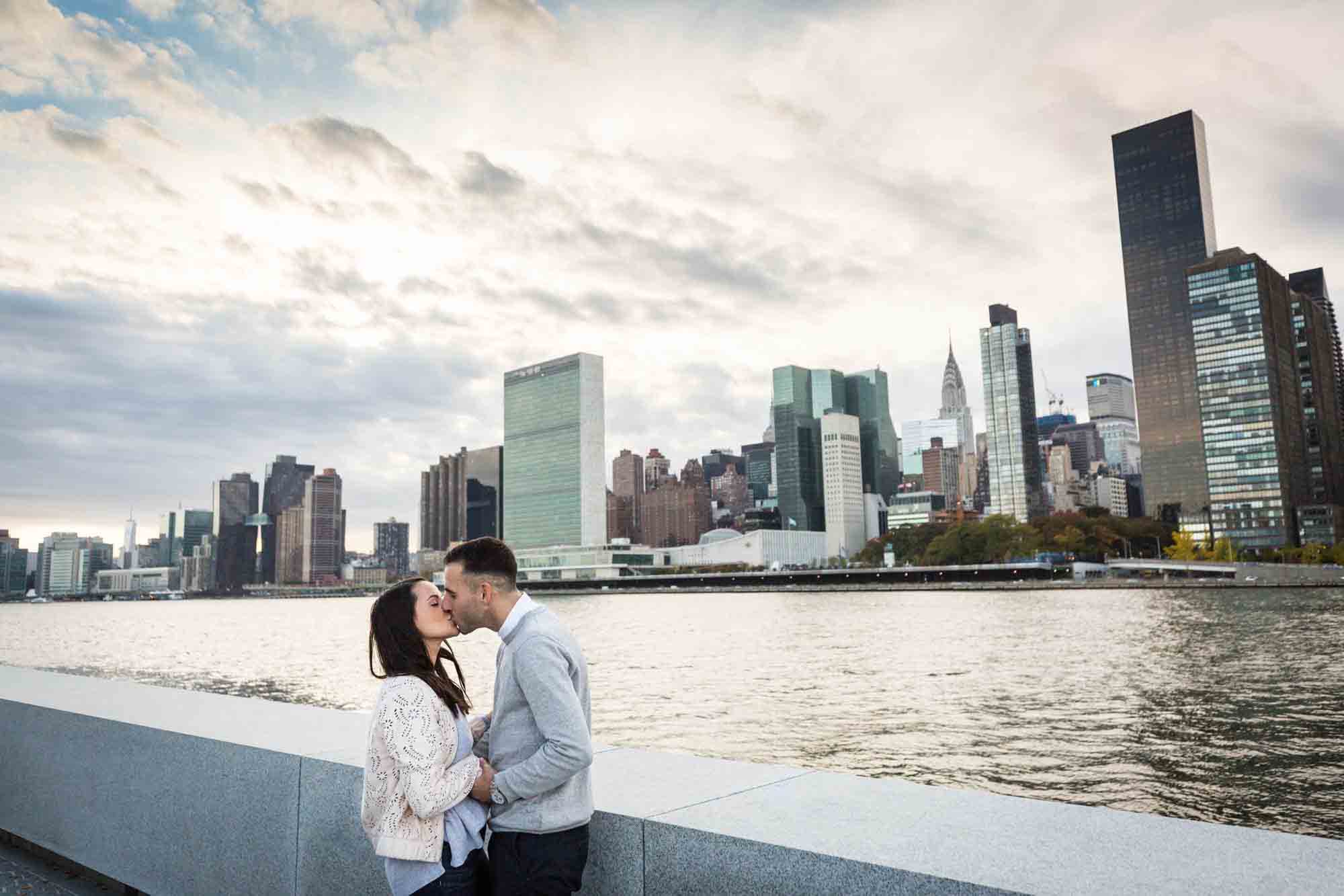 Couple kissing in front of NYC skyline during a Roosevelt Island engagement photo shoot