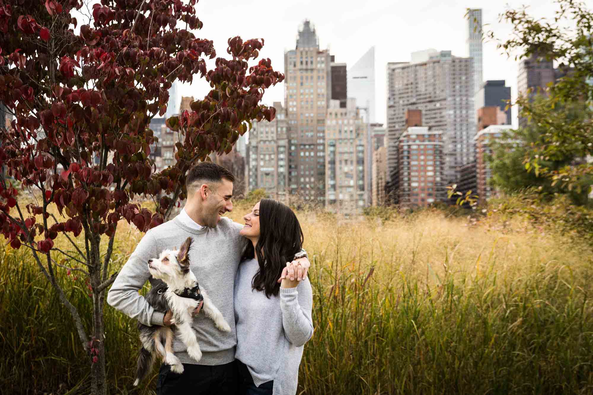 Couple hugging and holding dog in front of NYC skyline and grass for an article on pet engagement photo tips