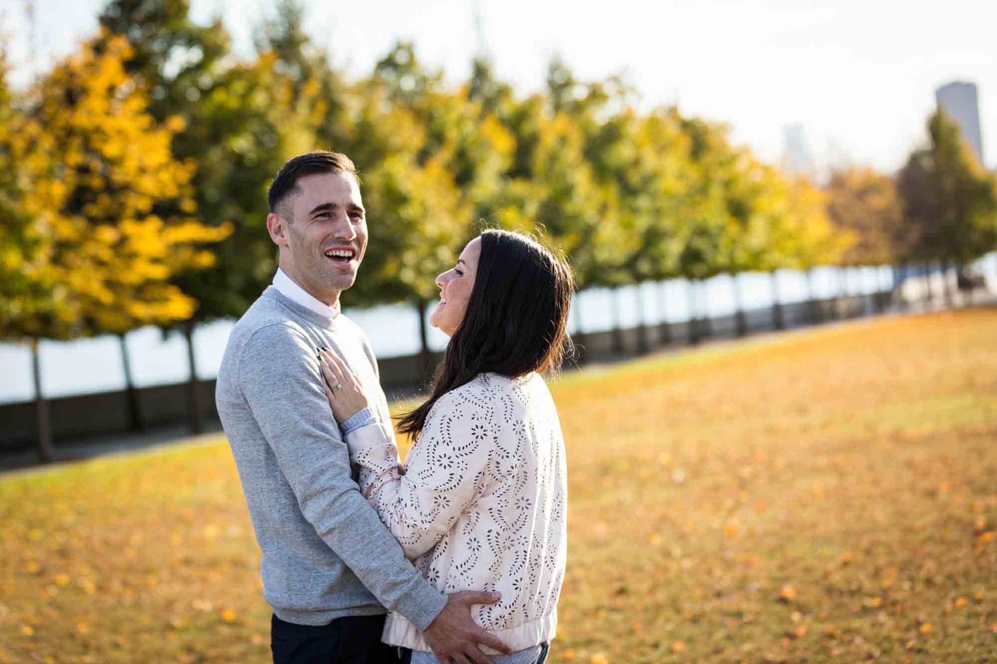 Couple laughing and hugging in front of grass and trees during a Roosevelt Island engagement photo shoot
