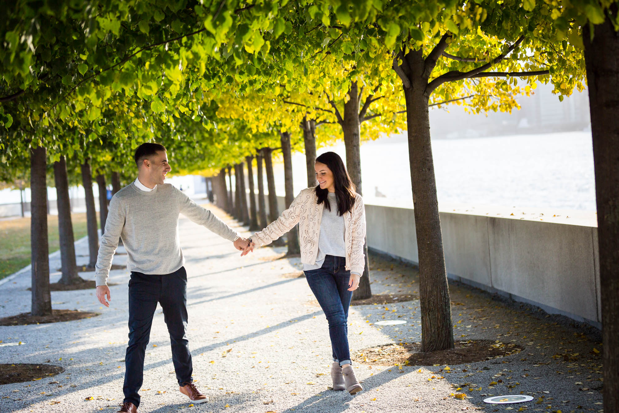 Couple holding hands and walking under trees during a Roosevelt Island engagement photo shoot