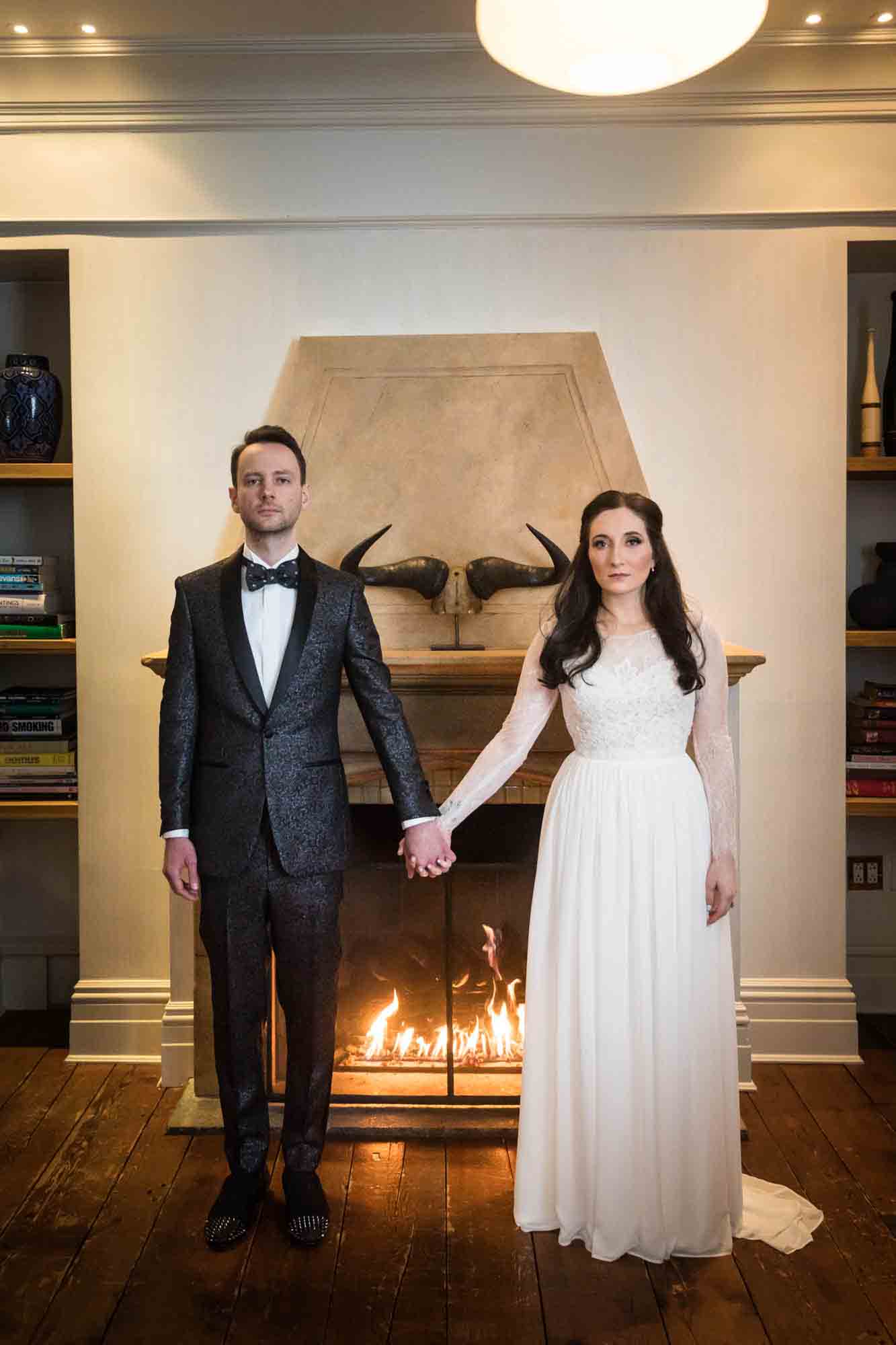 Bride and groom looking stern and holding hands in front of fireplace