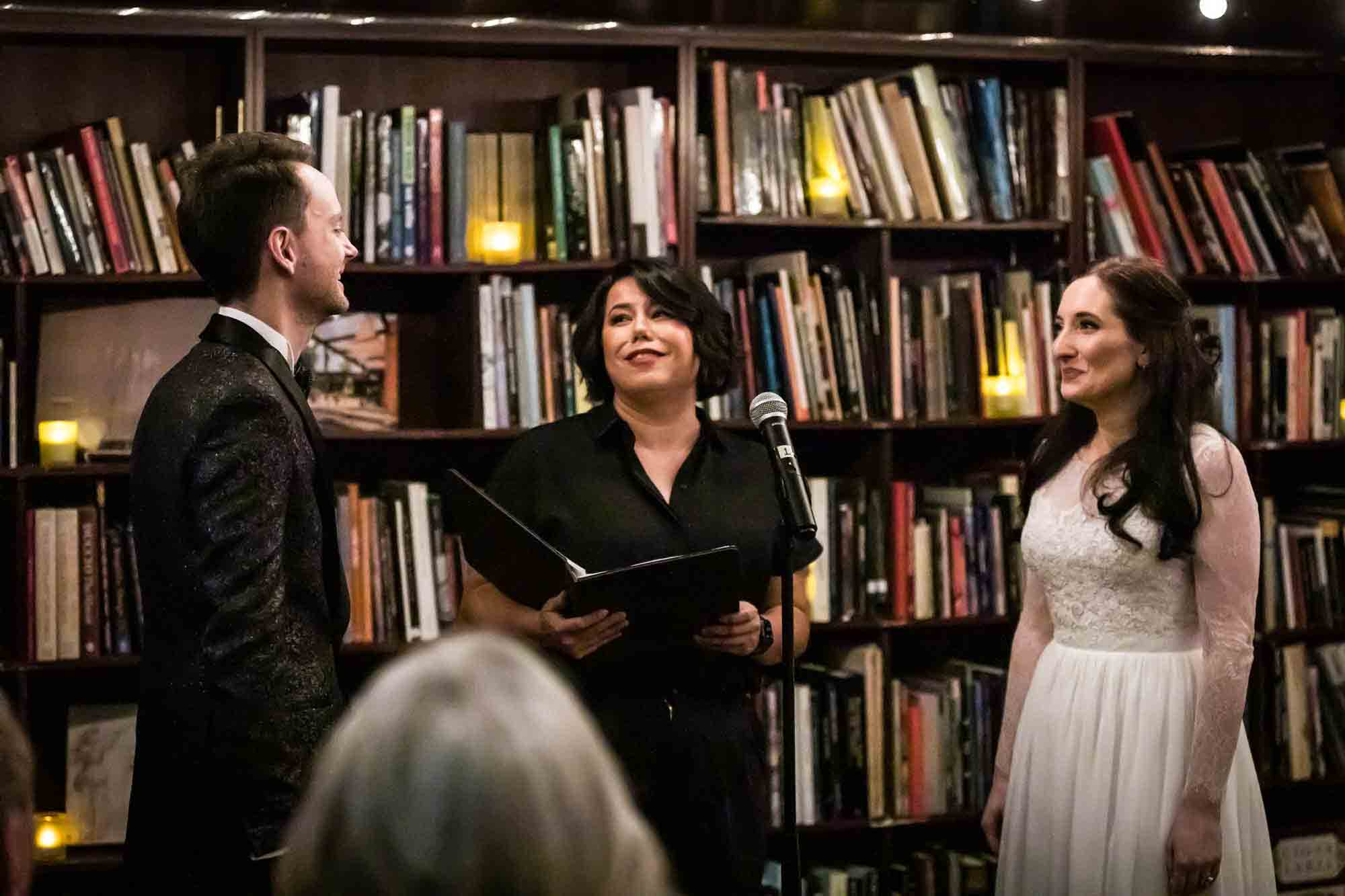 Bride and groom standing with officiant in bookstore for an article on wedding ceremony photo tips