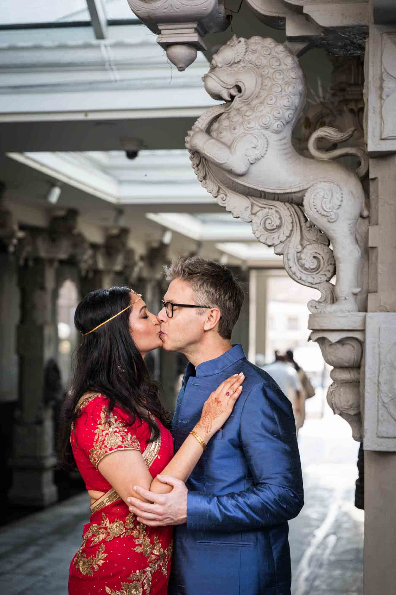 Ganesha Temple wedding photos of bride and groom kissing underneath elaborate stone pillar