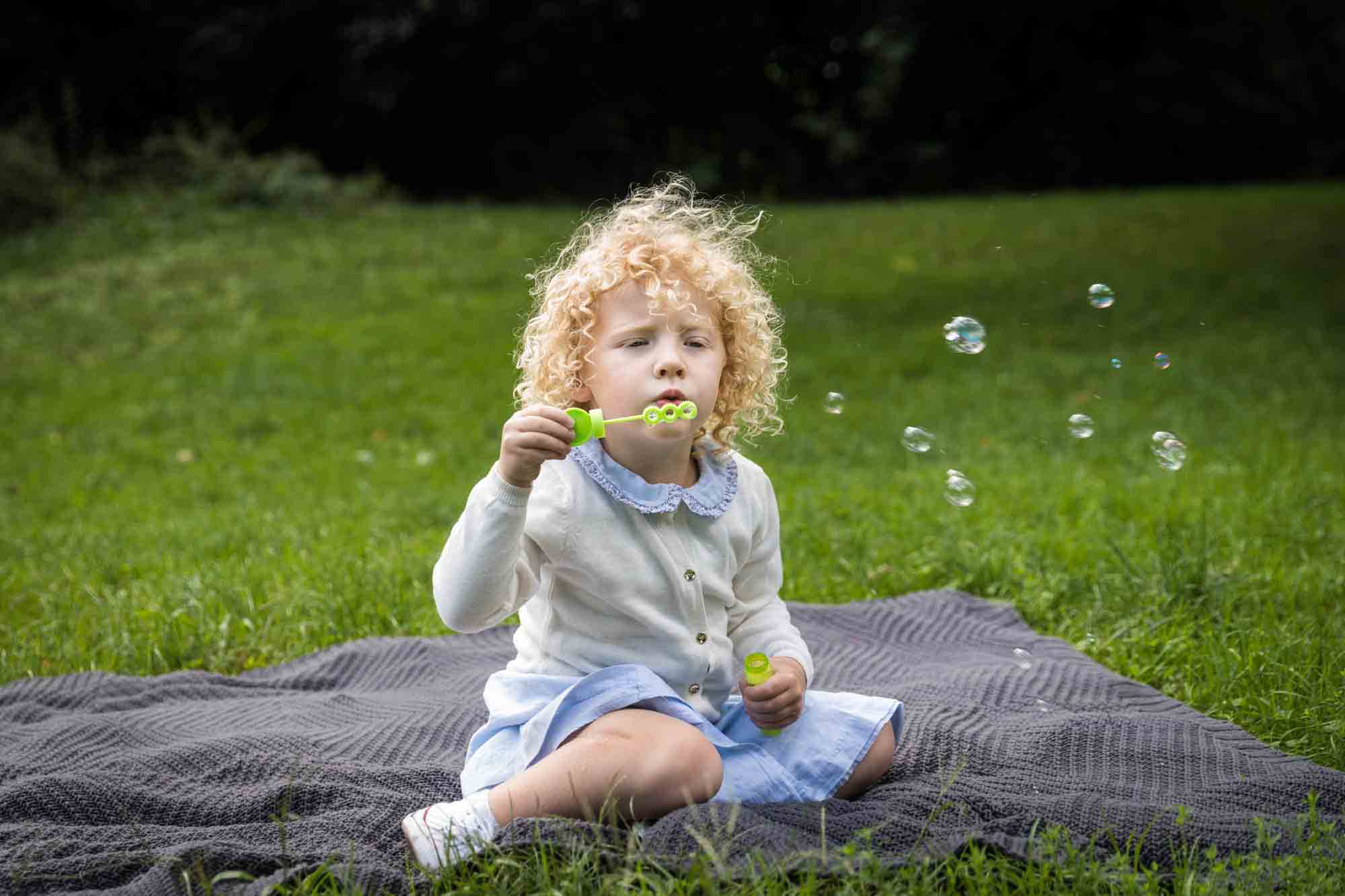 Little blonde girl sitting on blanket blowing bubbles for an article on NYC family portrait tips for tourists