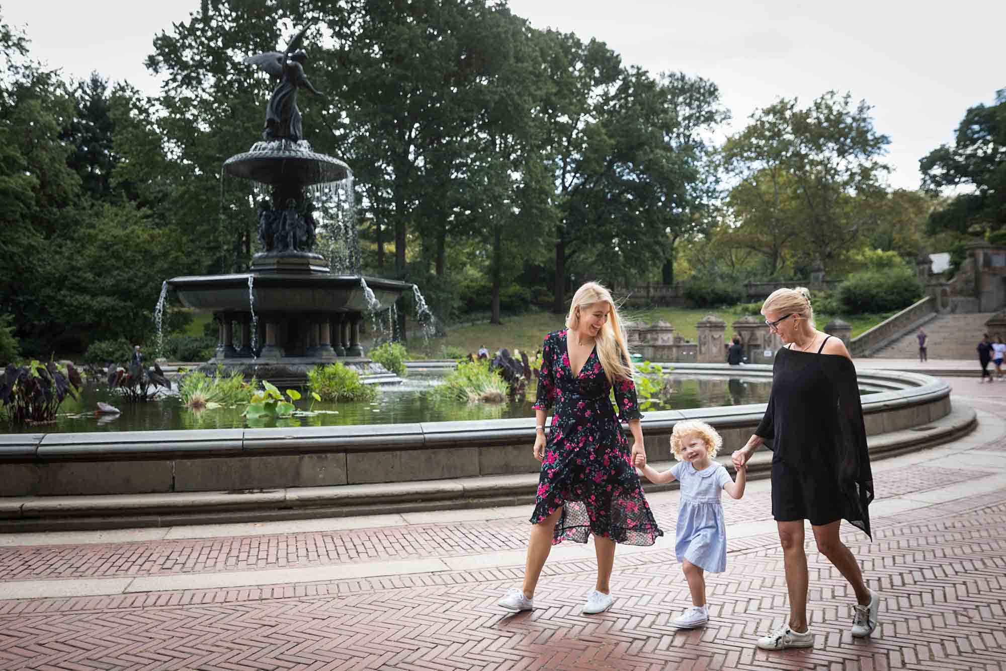 Mother and grandmother holding hands with little girl walking in front of Bethesda Fountain for an article on NYC family portrait tips for tourists