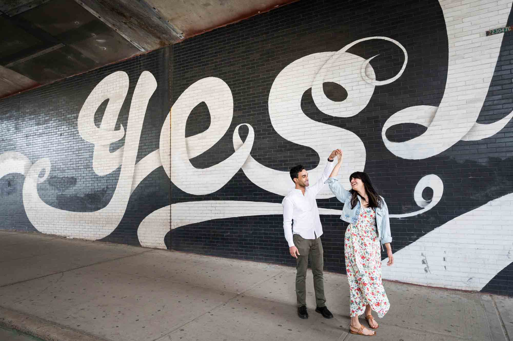 Couple dancing in front of black and white 'Yes' mural during a Brooklyn Bridge Park engagement photo shoot