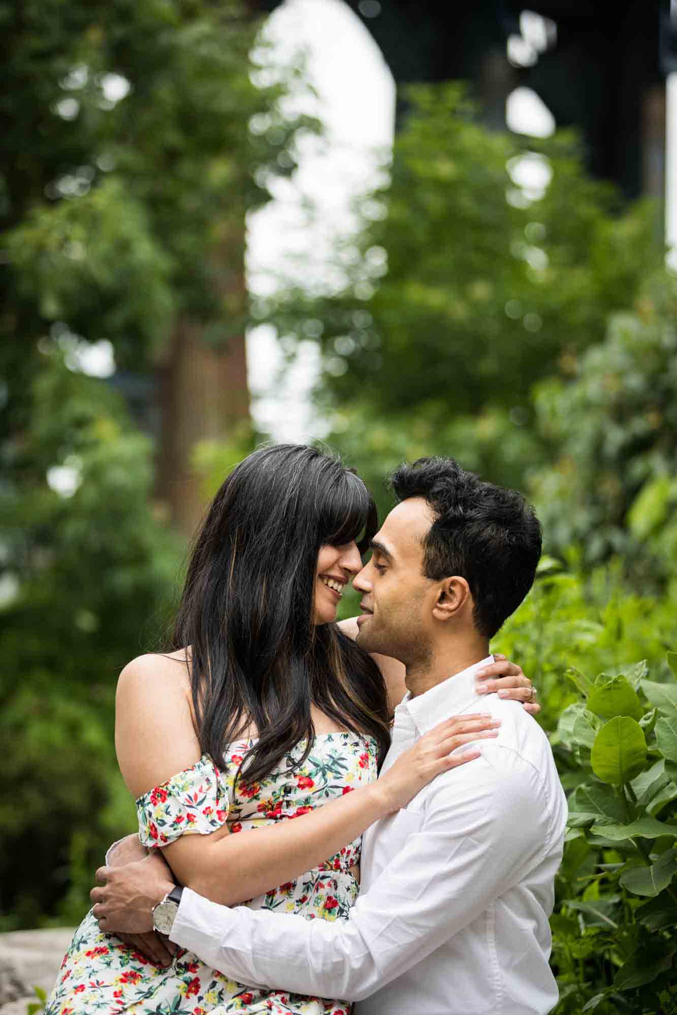 Woman sitting in man's lap in front of trees and bushes during a Brooklyn Bridge Park engagement photo shoot