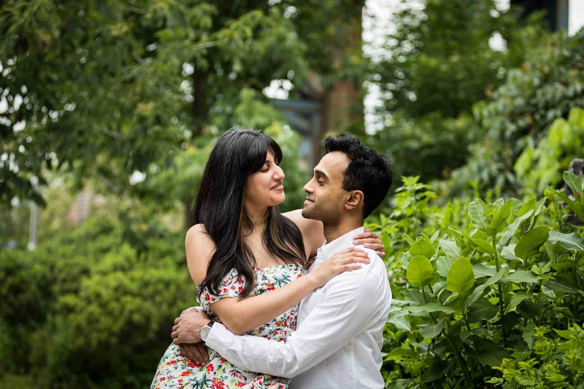 Woman sitting in man's lap in front of trees and bushes during a Brooklyn Bridge Park engagement photo shoot