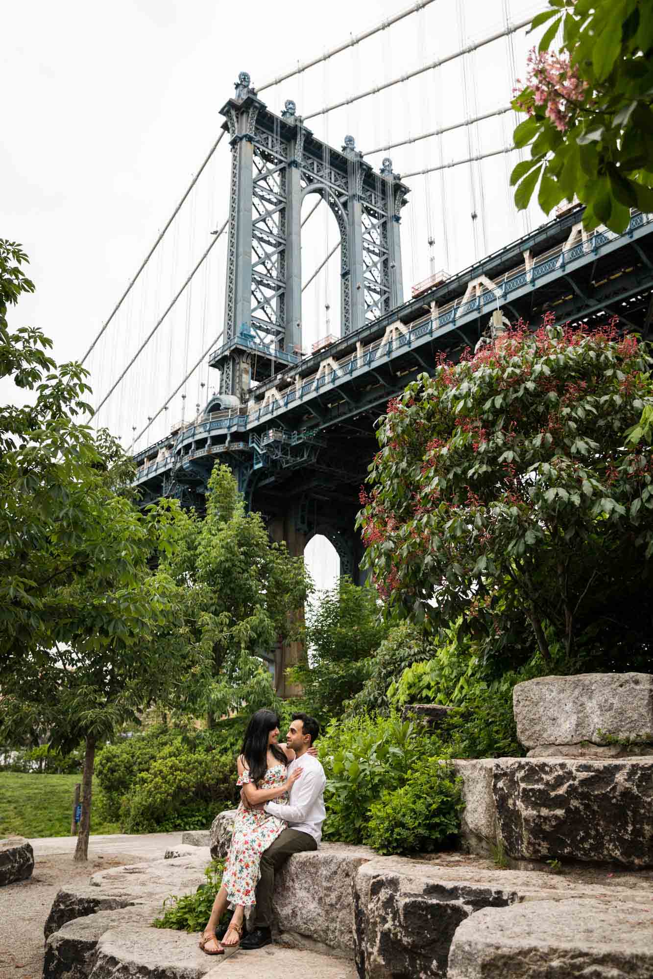 Woman sitting in man's lap in front of Manhattan Bridge during a Brooklyn Bridge Park engagement photo shoot