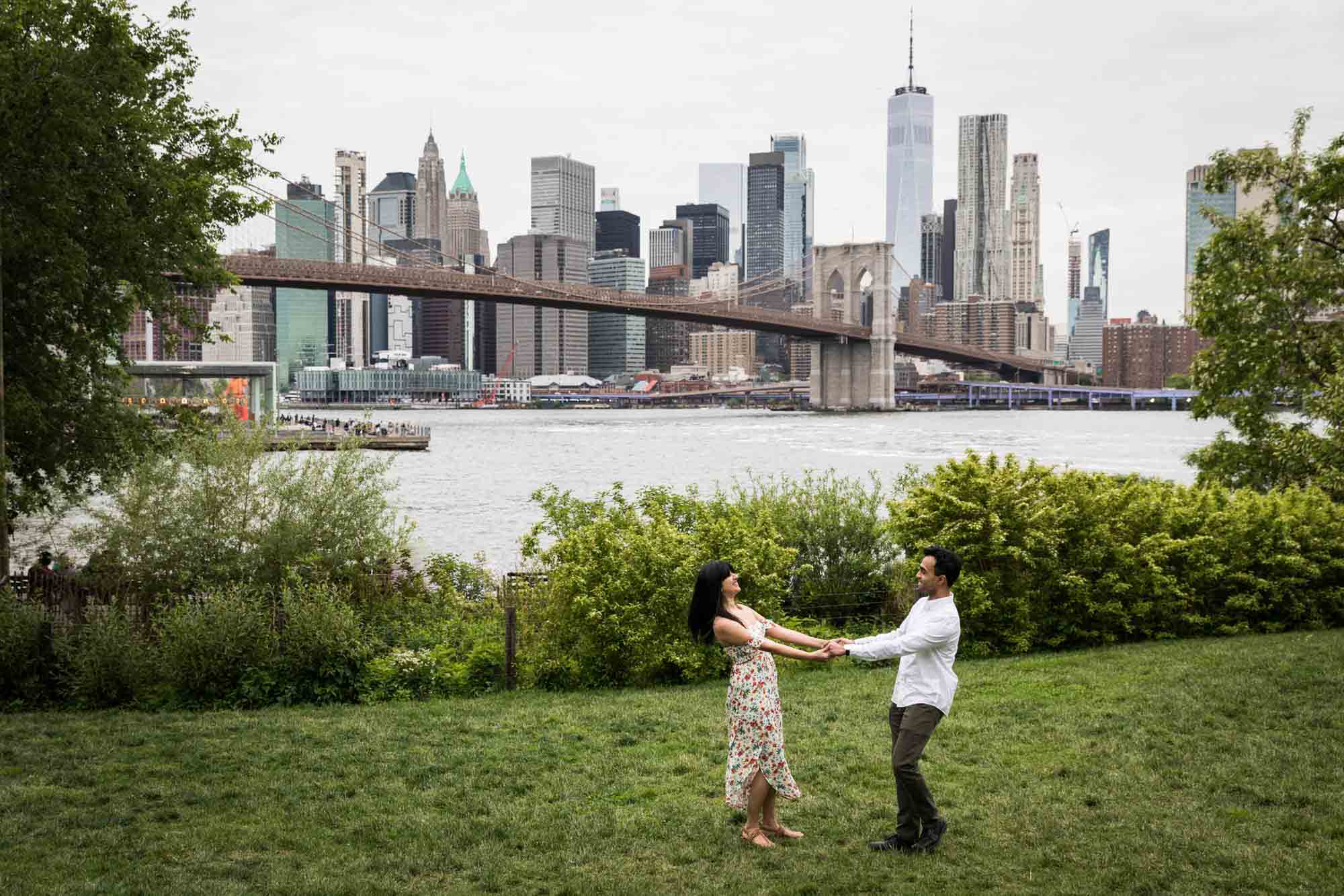 Couple dancing on lawn with view of NYC skyline behind during a Brooklyn Bridge Park engagement photo shoot