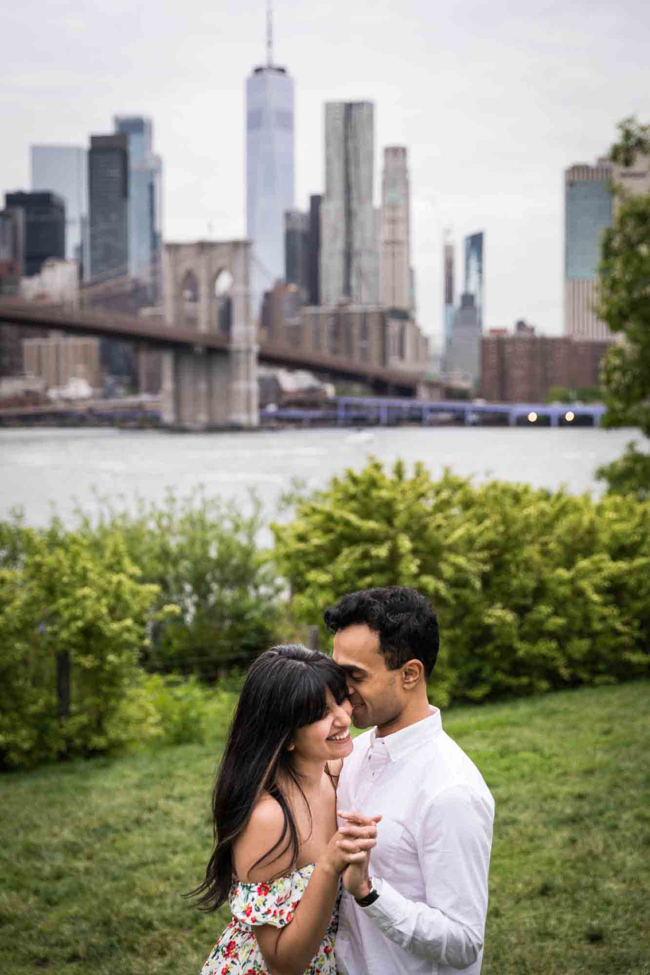 Couple dancing in grass with NYC skyline in background during a Brooklyn Bridge Park engagement photo shoot