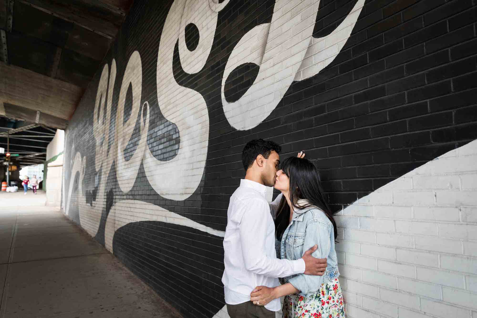 Couple kissing against wall in front of 'Yes' mural during a Brooklyn Bridge Park engagement photo shoot