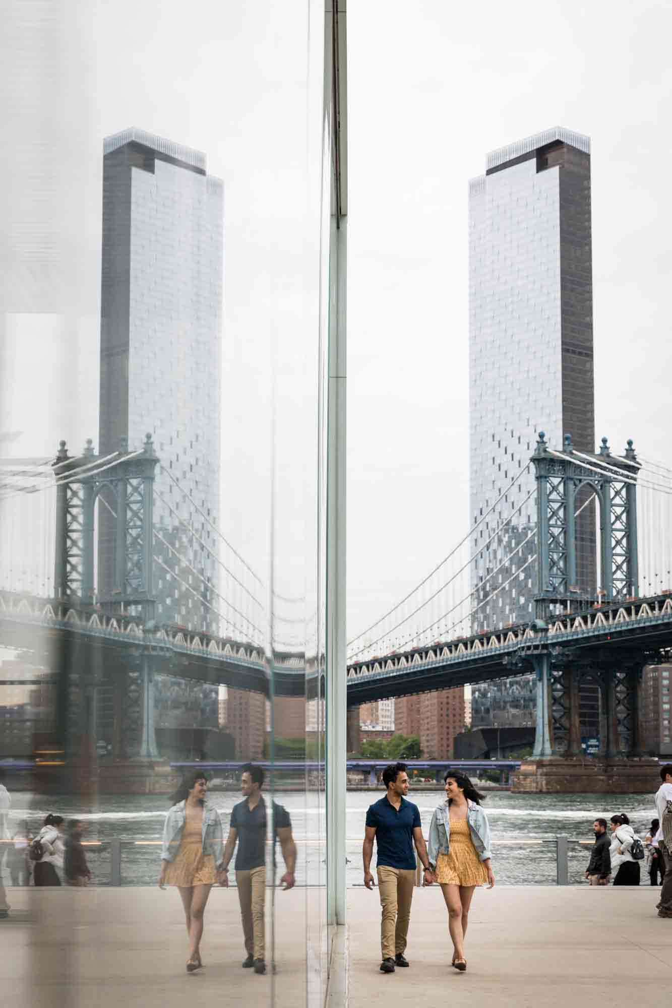 Brooklyn Bridge Park engagement photo of couple walking beside Jane's Carousel with reflection in glass