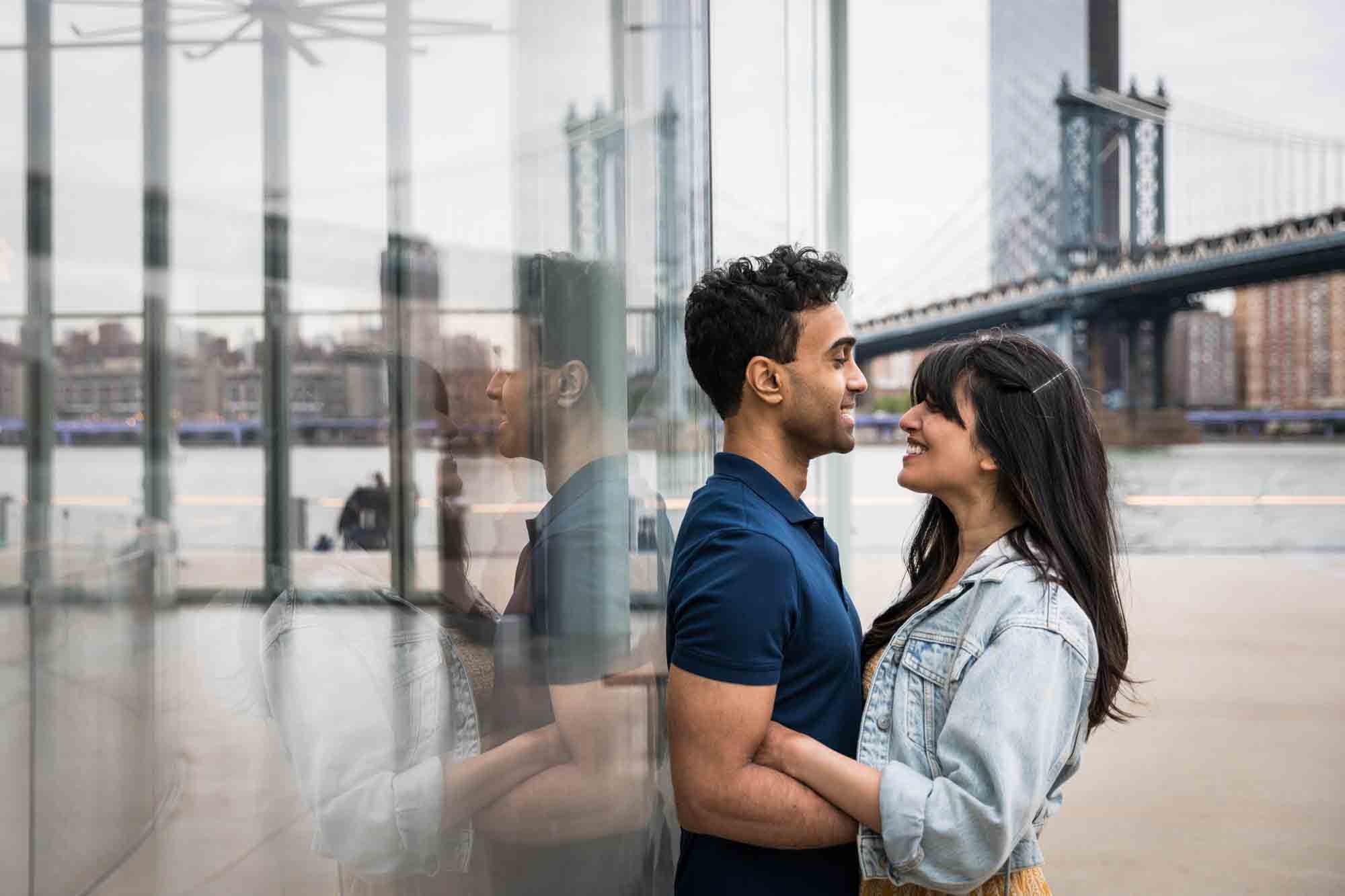 Brooklyn Bridge Park engagement photo of couple cuddling against glass wall of Jane's Carousel