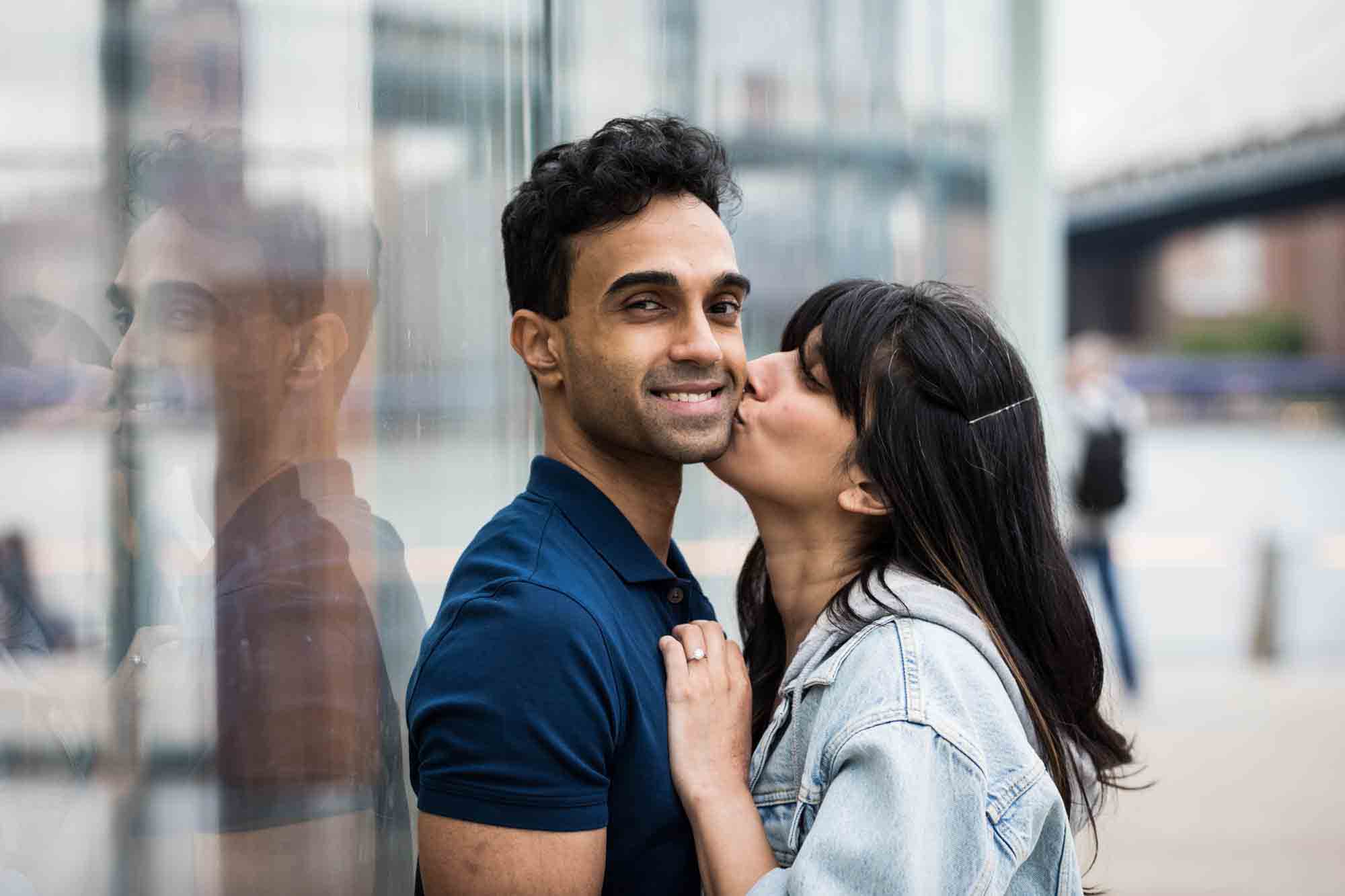 Brooklyn Bridge Park engagement photo of woman kissing man on the cheek against glass wall