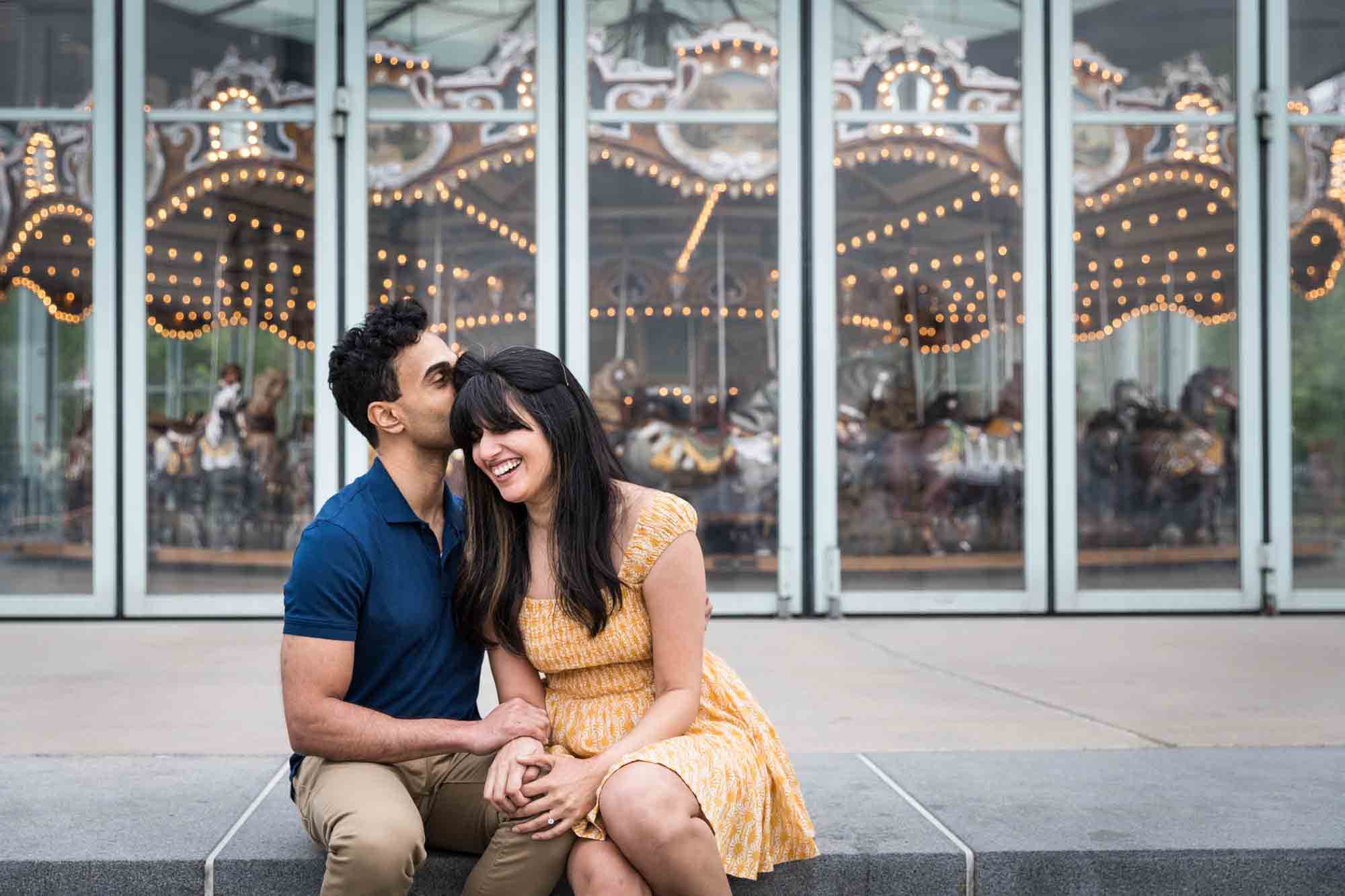 Man kissing woman on the head in front of Jane's Carousel during a Brooklyn Bridge Park engagement photo shoot