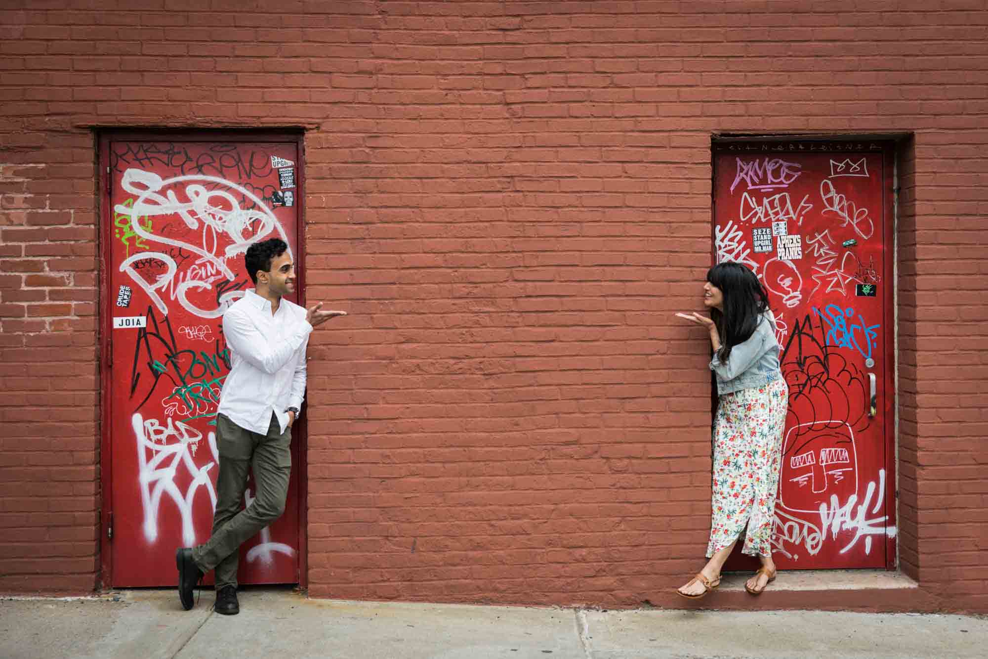 Couple blowing each other kisses in front of red doors during a Brooklyn Bridge Park engagement photo shoot